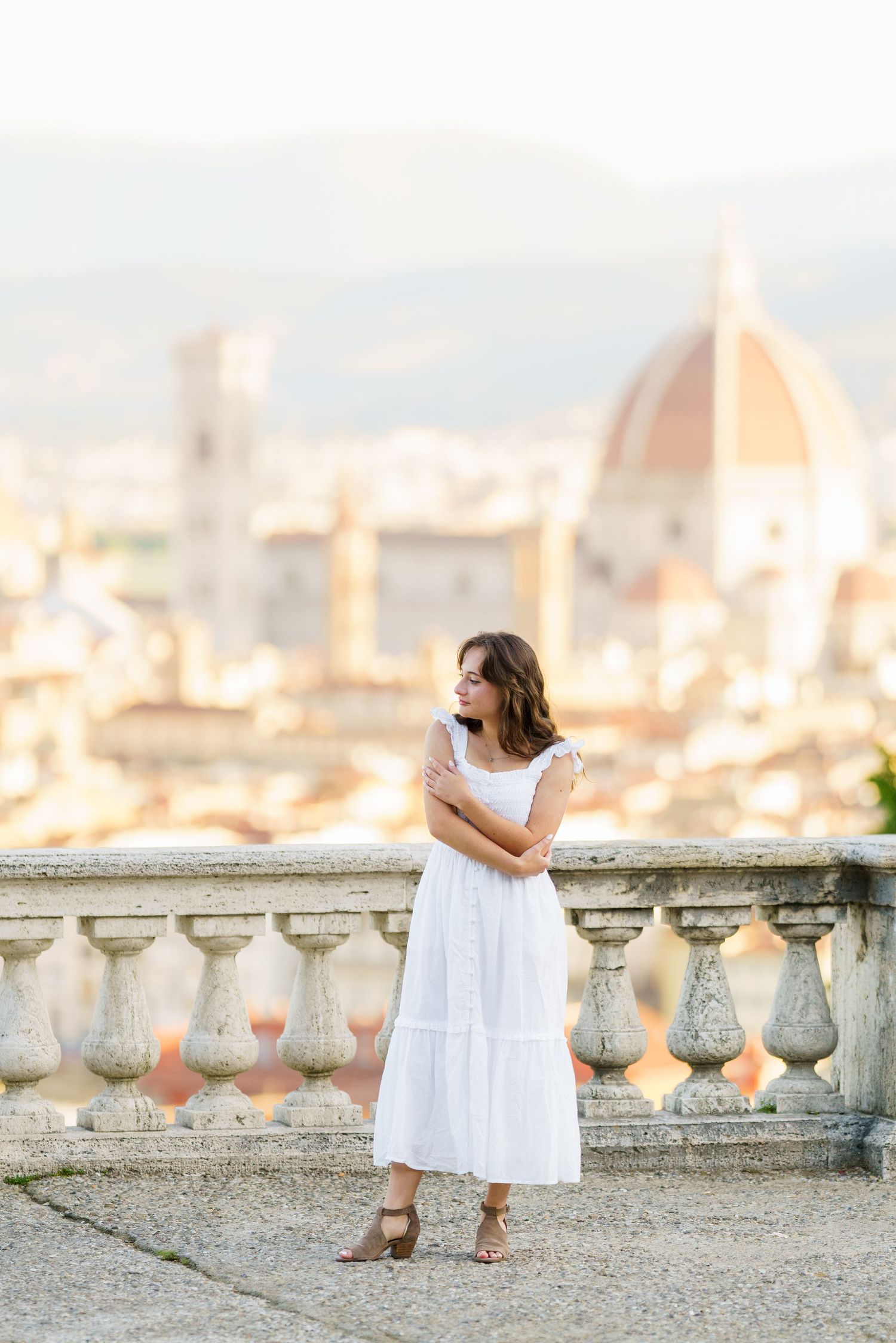 Two figures in white dresses stand together on a stone terrace overlooking a scenic city view at sunset.