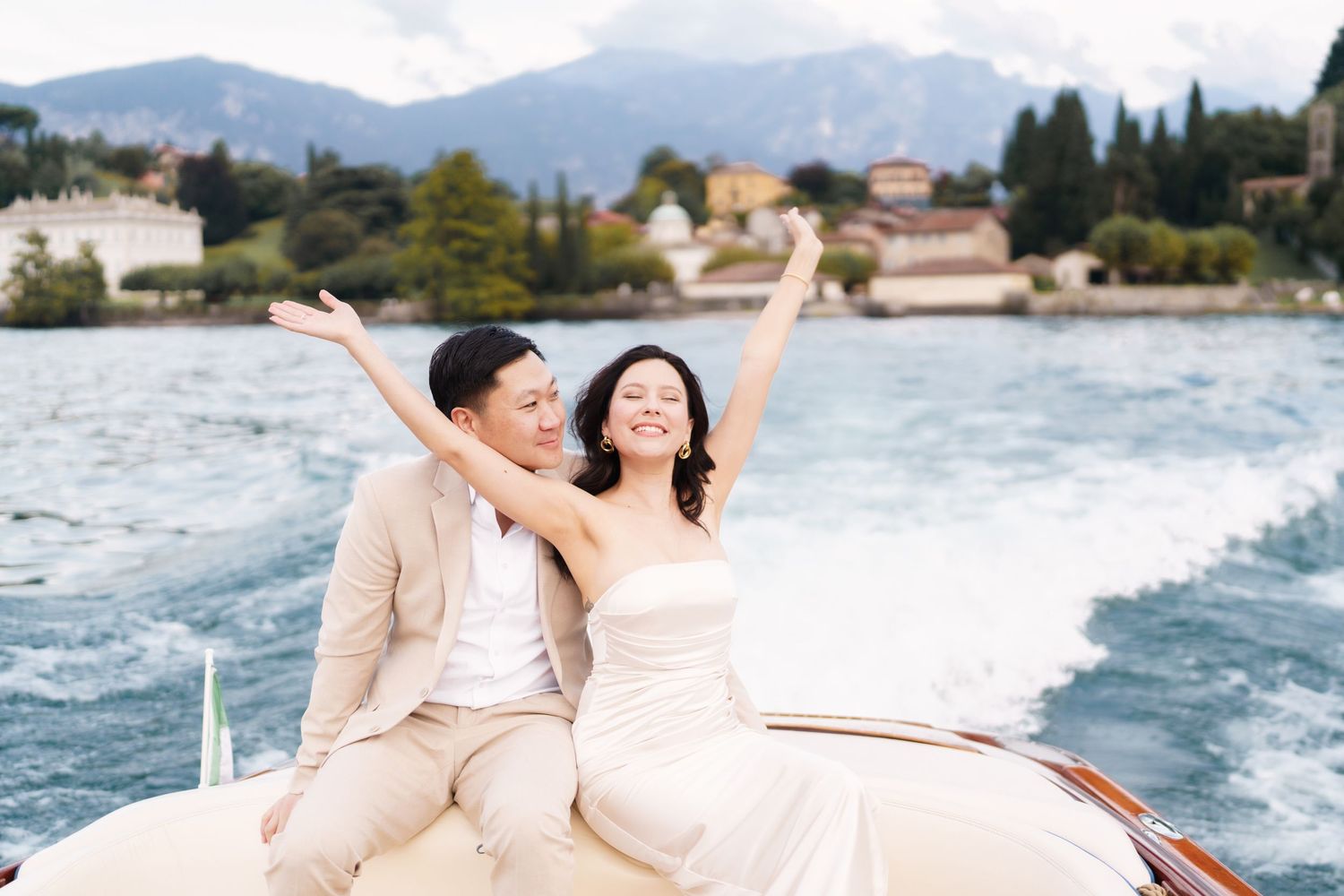 A couple celebrates joyfully while riding on a speedboat across sparkling waters with mountains in the background.