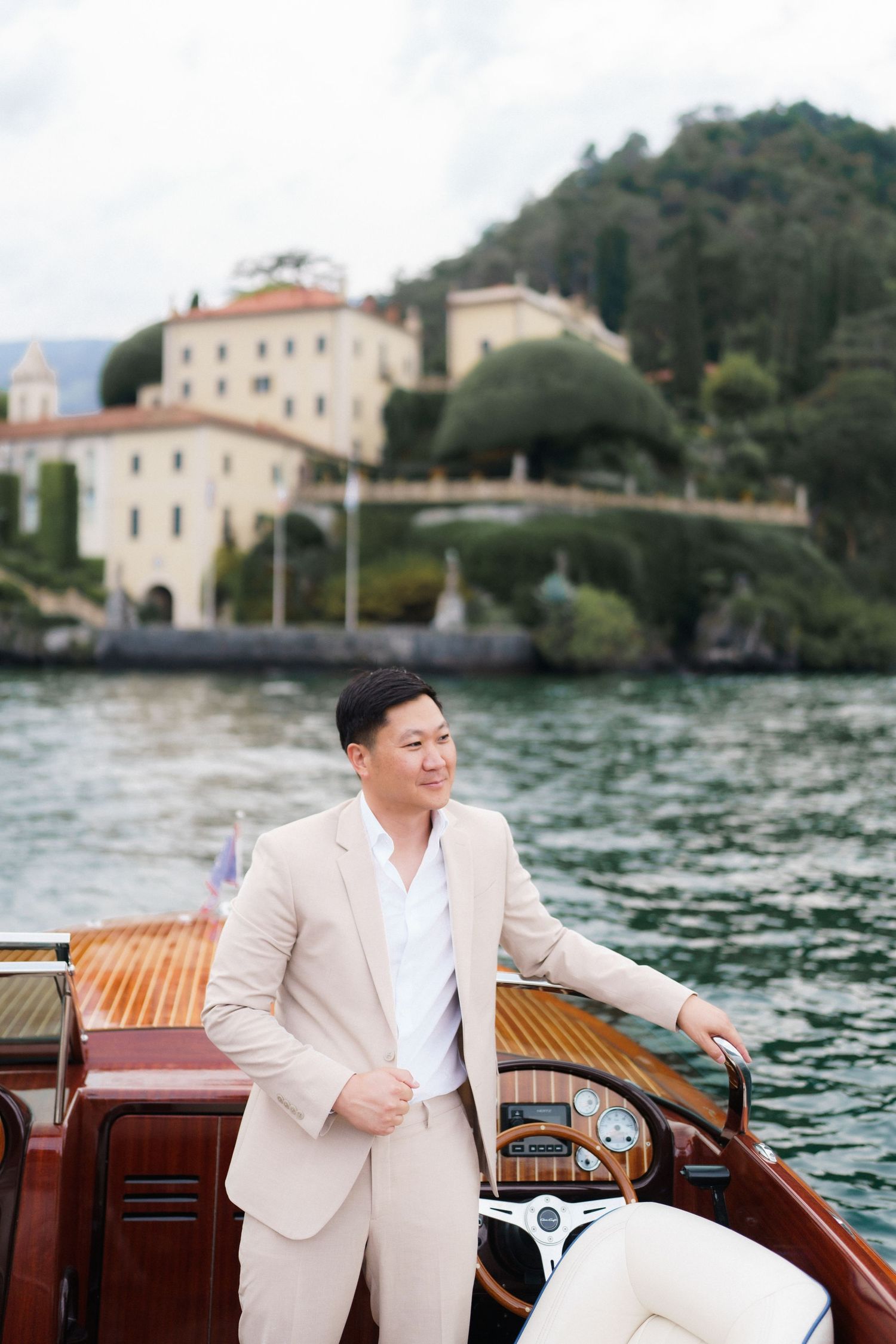 Person in beige suit standing on wooden boat with Italian coastal villa and blue water in background.