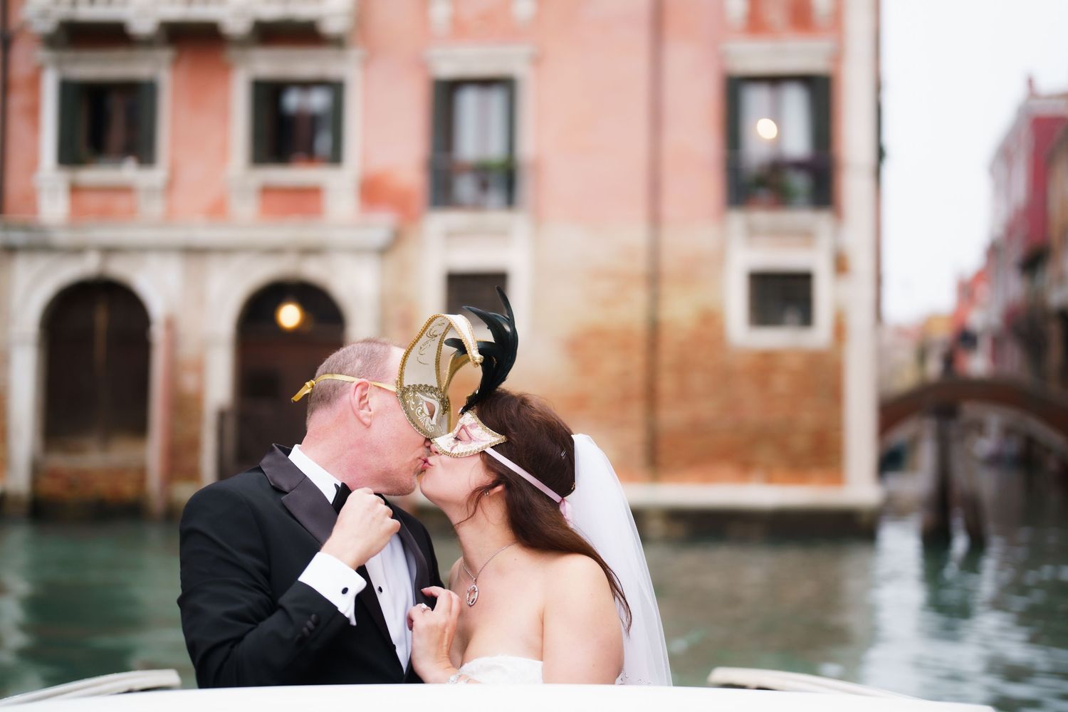 A romantic wedding couple shares a kiss while standing on a bridge in Venice with historic buildings and canals in view.