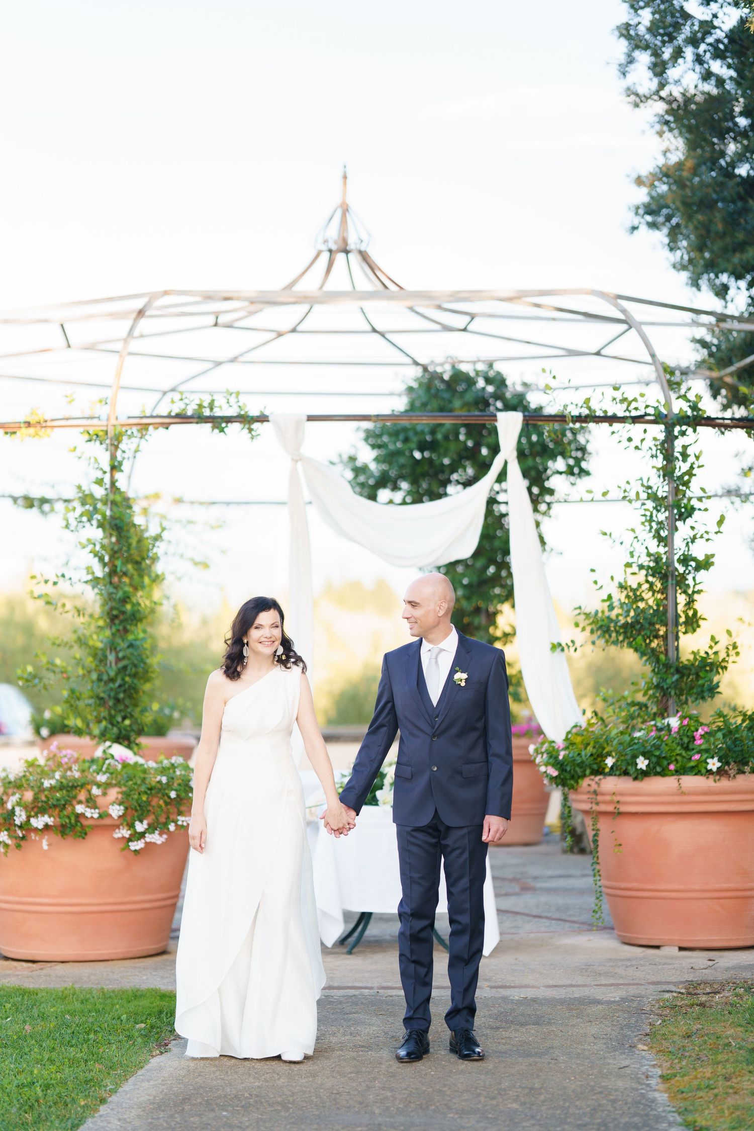 Elegant wedding couple strolling along outdoor pathway surrounded by terracotta planters.