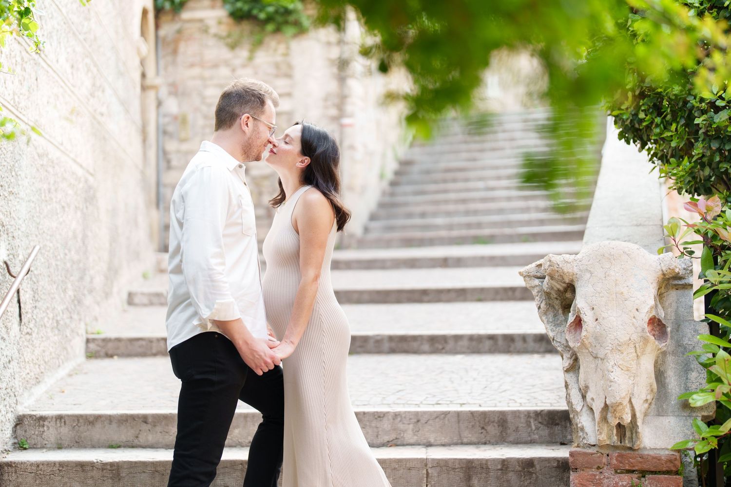 A couple shares a romantic kiss on stone steps surrounded by lush greenery and historic architecture.