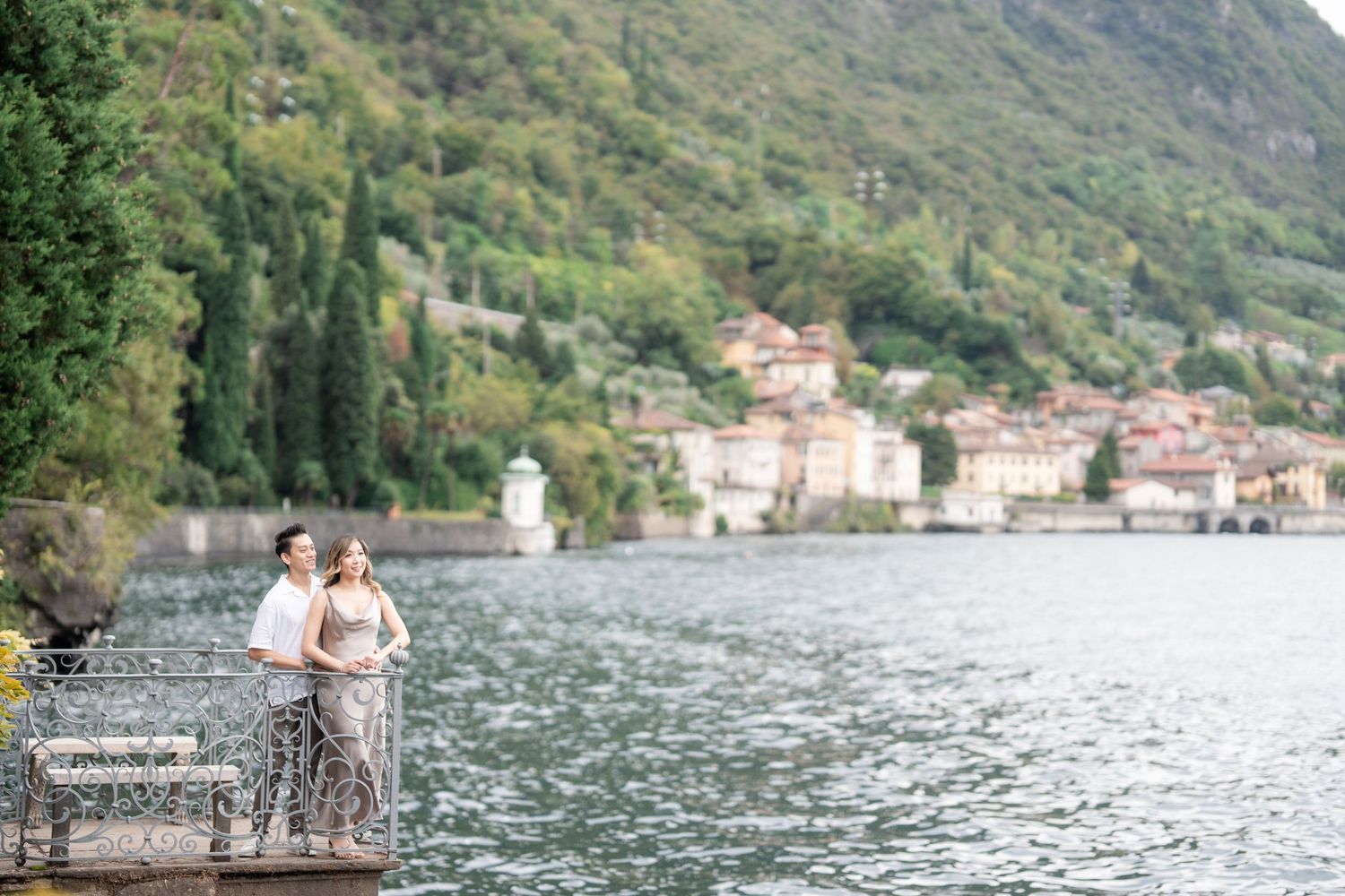 Scenic view of Lake Como with a couple embracing on a waterfront terrace overlooking Italian lakeside village.