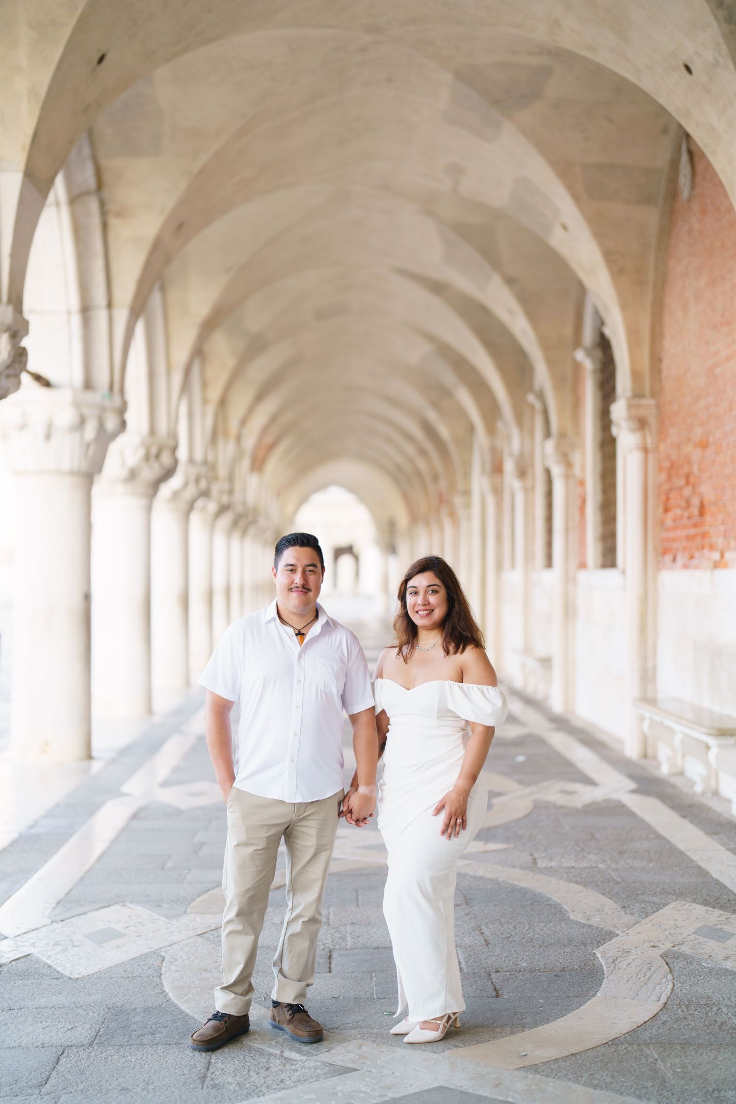 A couple in white attire walks through an elegant arched colonnade with repeating architectural columns and shadows.