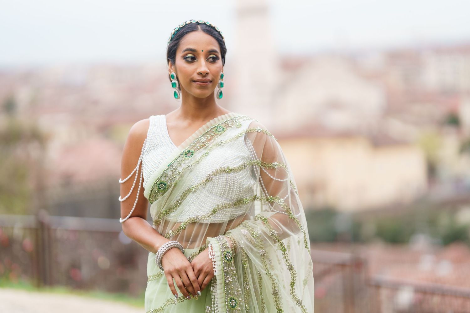 A person wearing a light green and gold saree poses outdoors against a blurred landscape background.