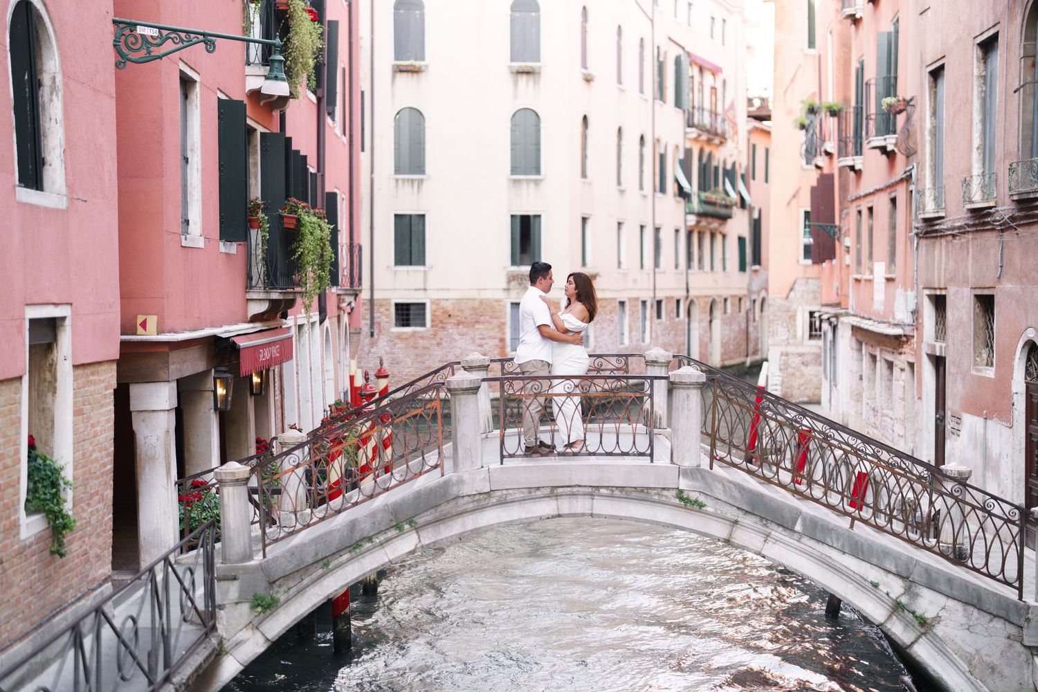 Romantic scene on a small bridge over a Venetian canal with pink buildings and ornate railings in Venice, Italy.