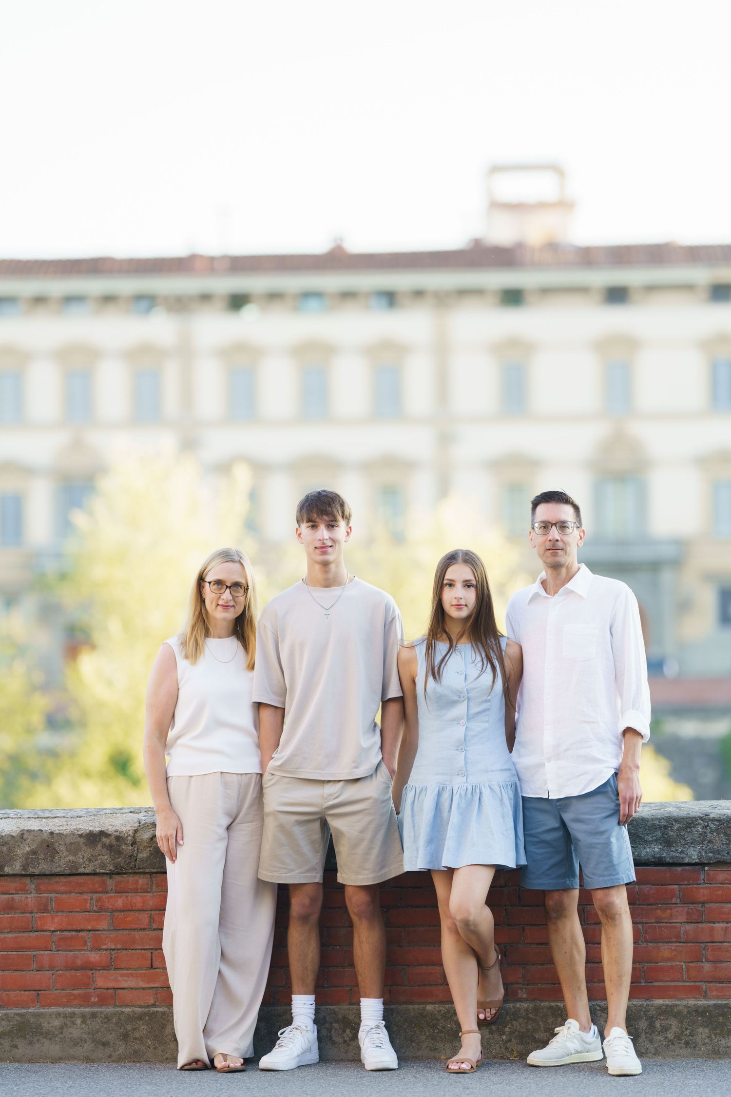 A family wearing coordinated white and beige outfits poses together on a brick wall at sunset.