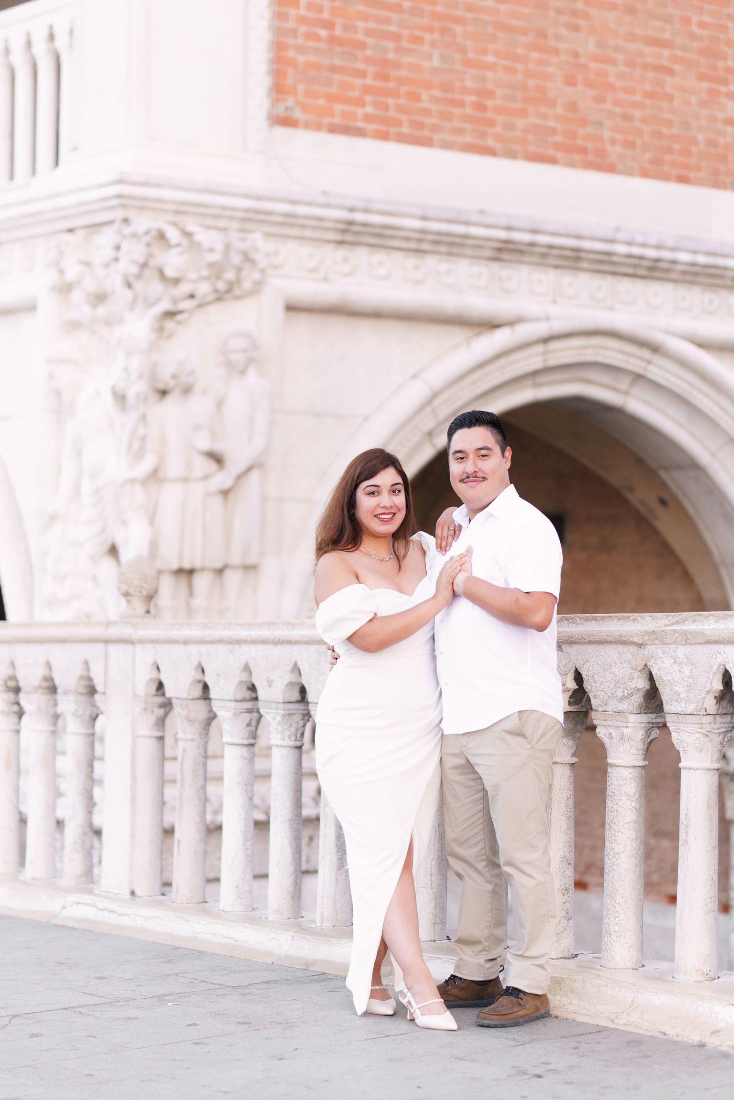 A couple in white outfits poses together against an ornate stone archway and columns of a historic building.
