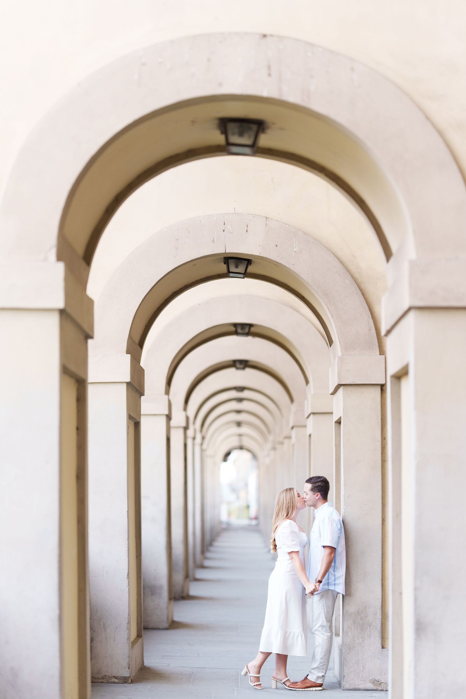 A couple stands in an elegant archway corridor with repeating white arches creating a beautiful vanishing point perspective.