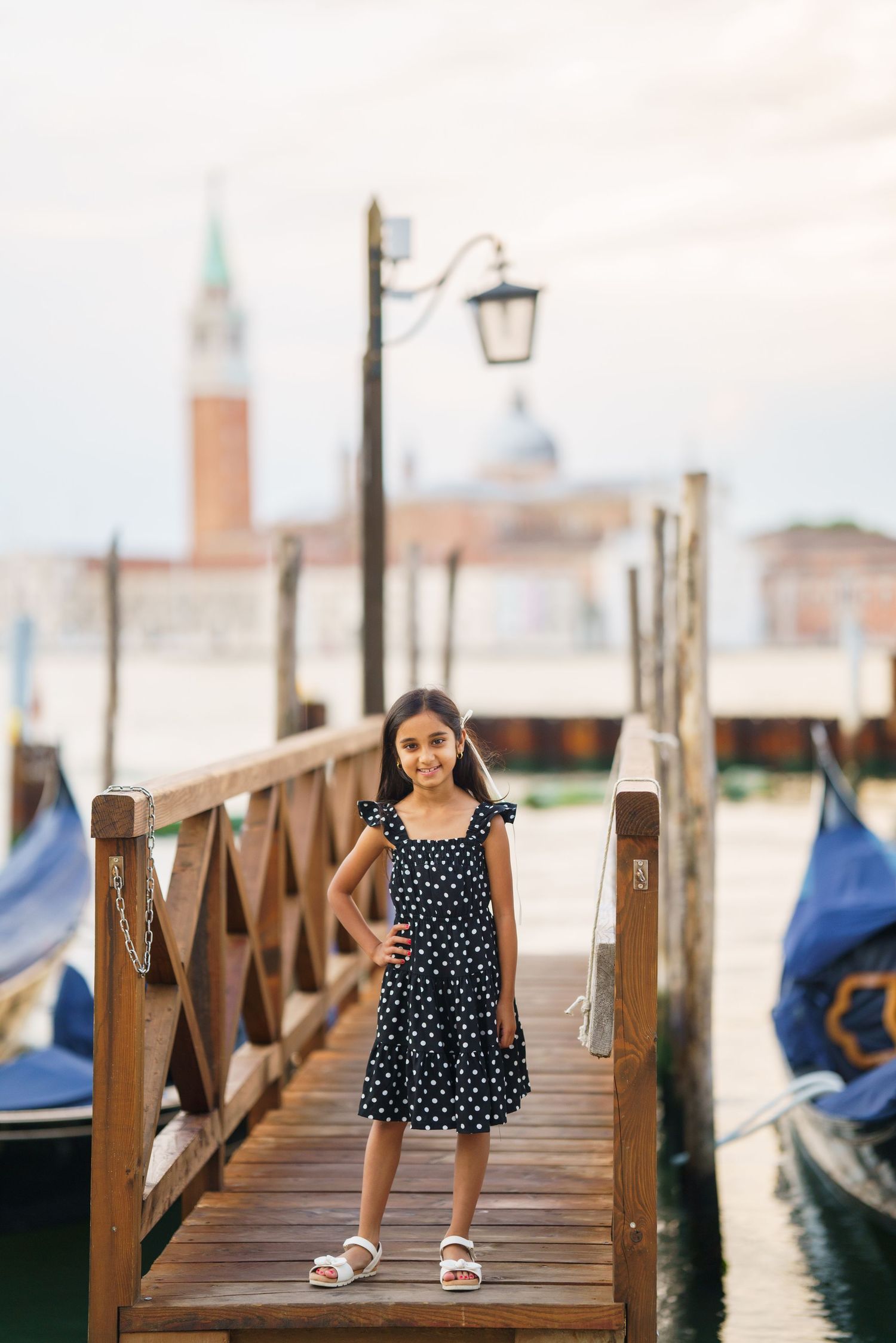 Young girl in polka dot dress posing on wooden dock in Venice, Italy, with gondolas and San Giorgio Maggiore in background.