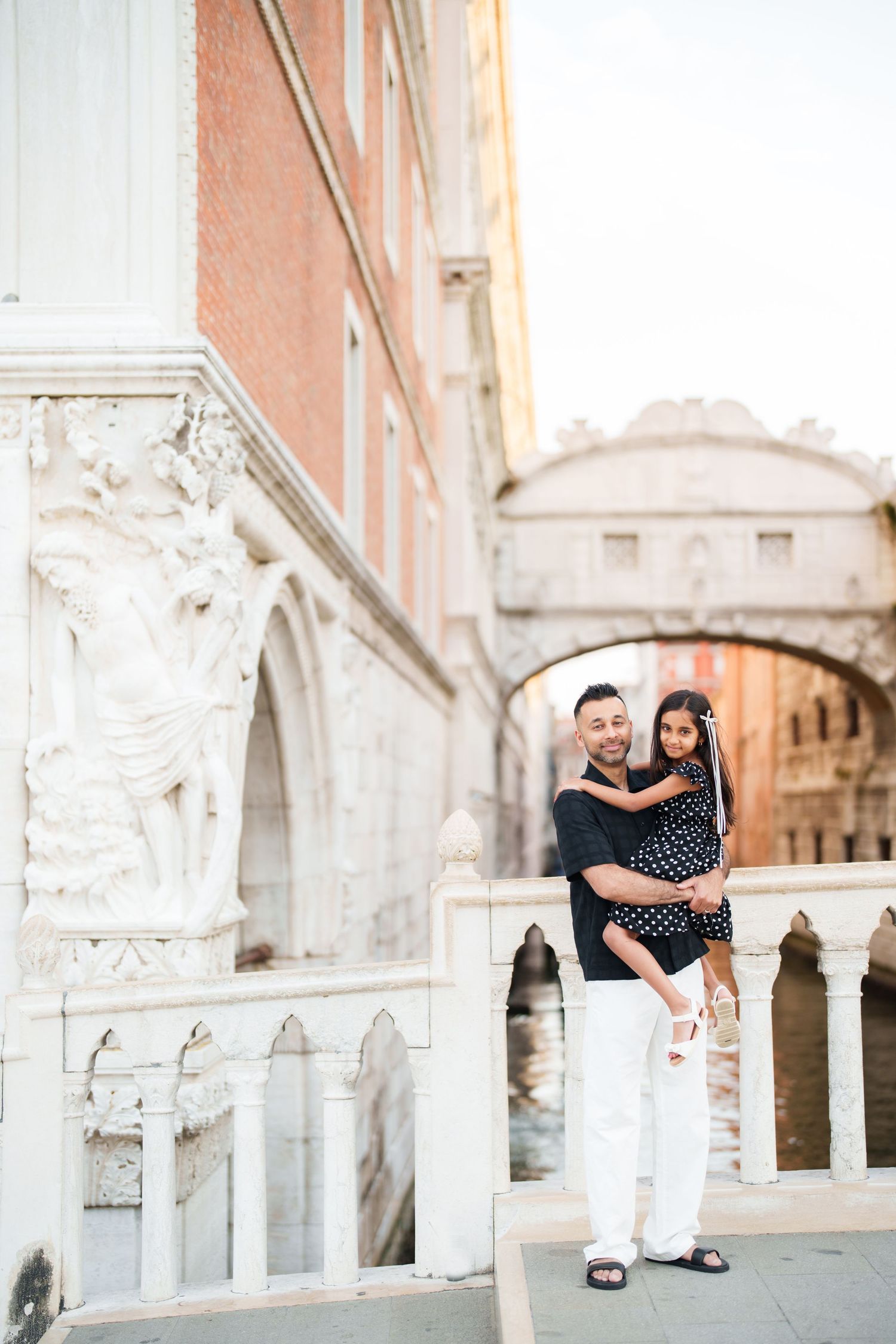 A couple embraces on a bridge near the Bridge of Sighs in Venice, Italy, with ornate white marble architecture.