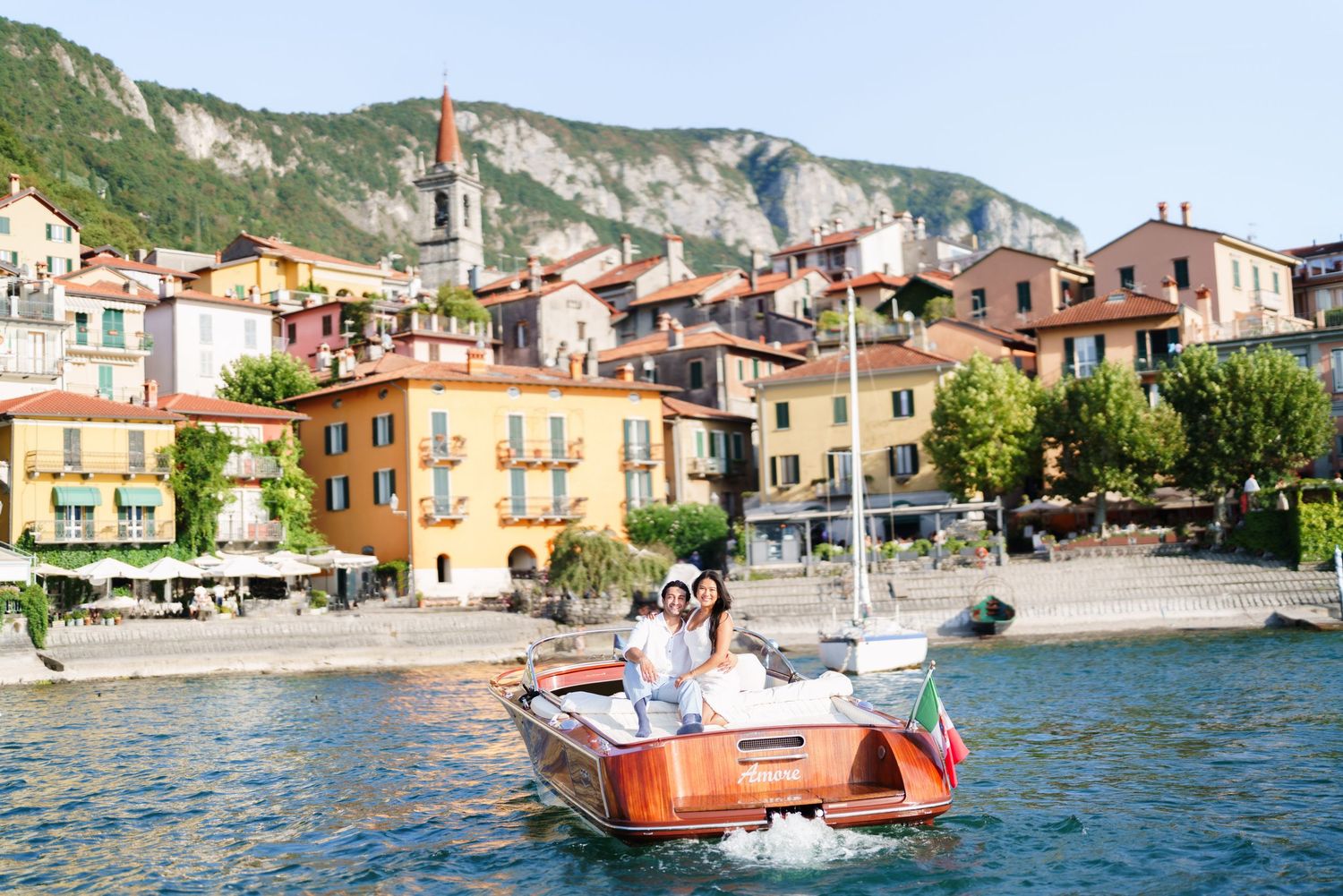 Couple sitting on a wooden boat on Lake Como with colorful Italian village buildings and mountains in the background.