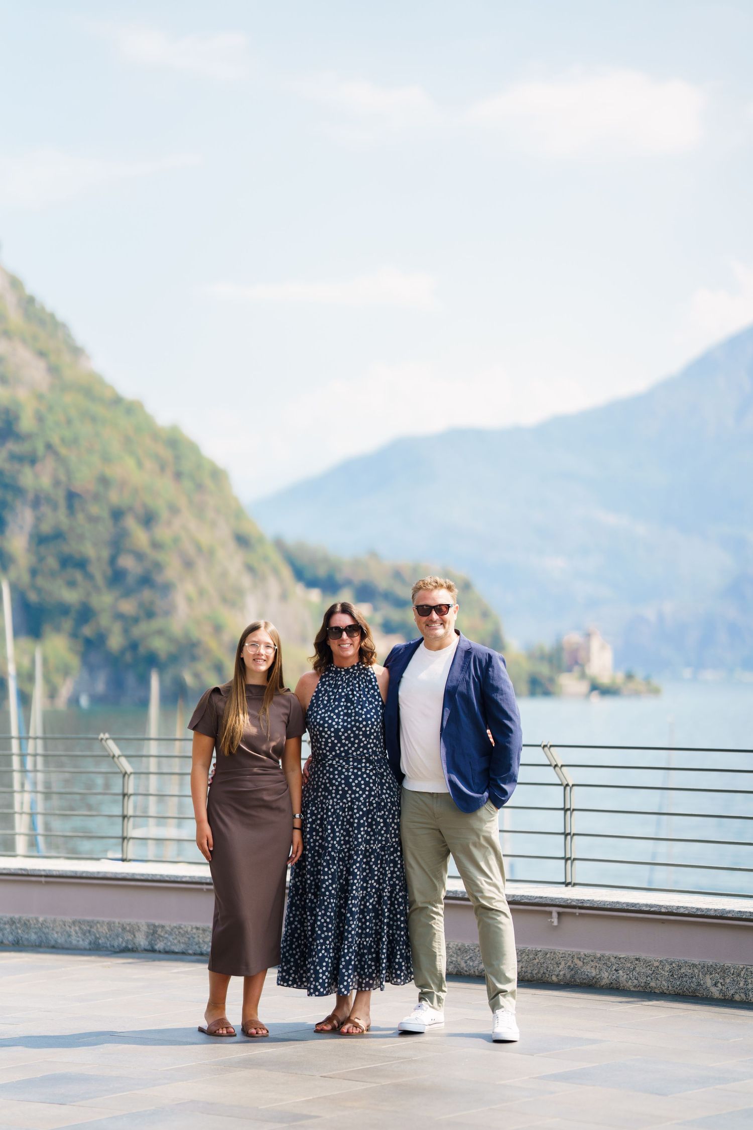 Three people posing on a lakeside promenade with scenic mountain and water views in the background.
