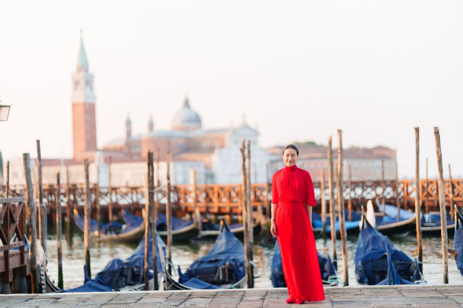 Woman in red dress posing by Venice gondolas with San Giorgio Maggiore church in background.