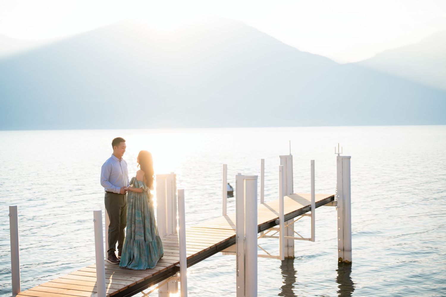 Couple standing on a wooden dock at sunset with calm lake and misty mountains in the background.