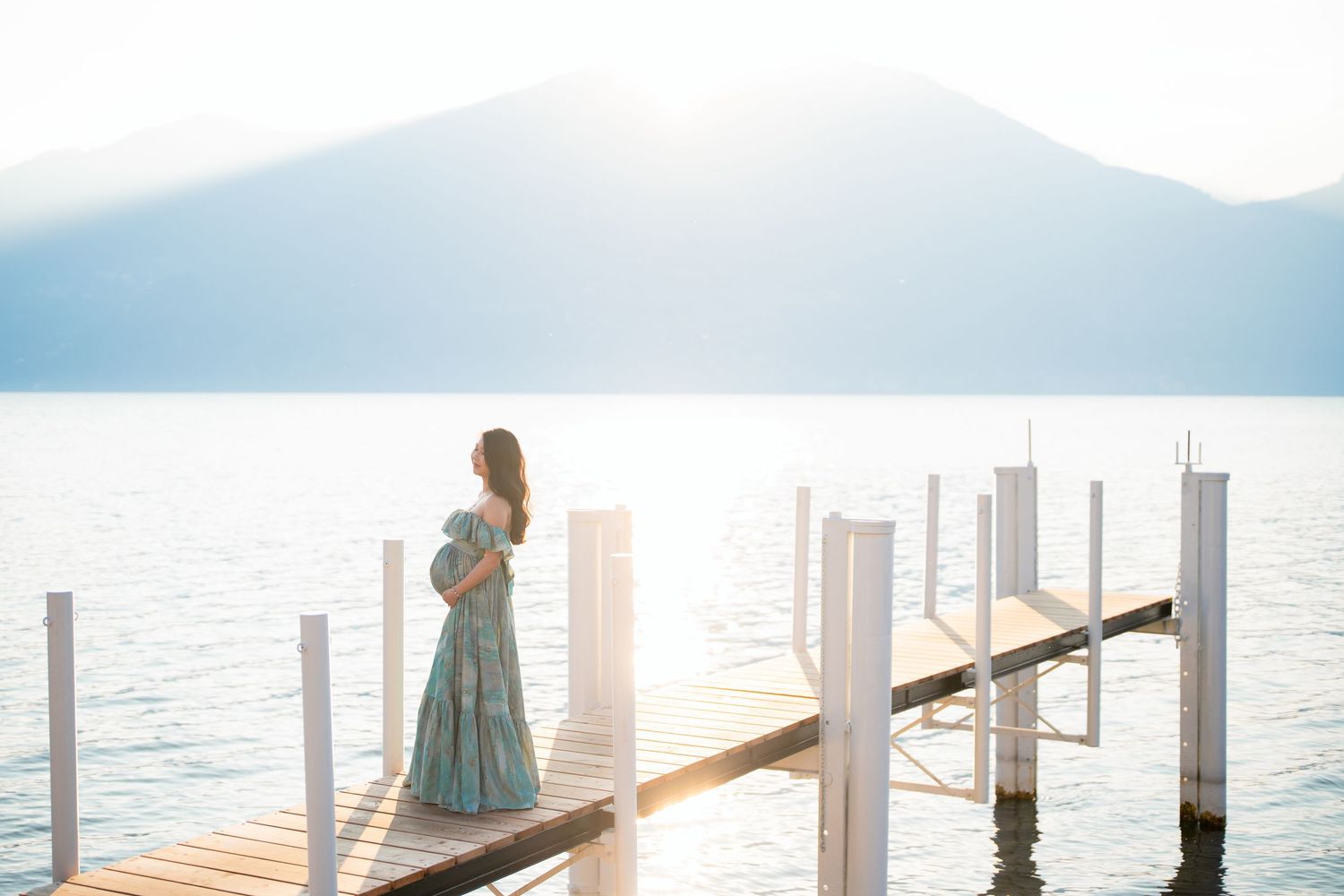 Pregnant woman in teal floral dress standing on wooden dock overlooking serene mountain lake at sunset.