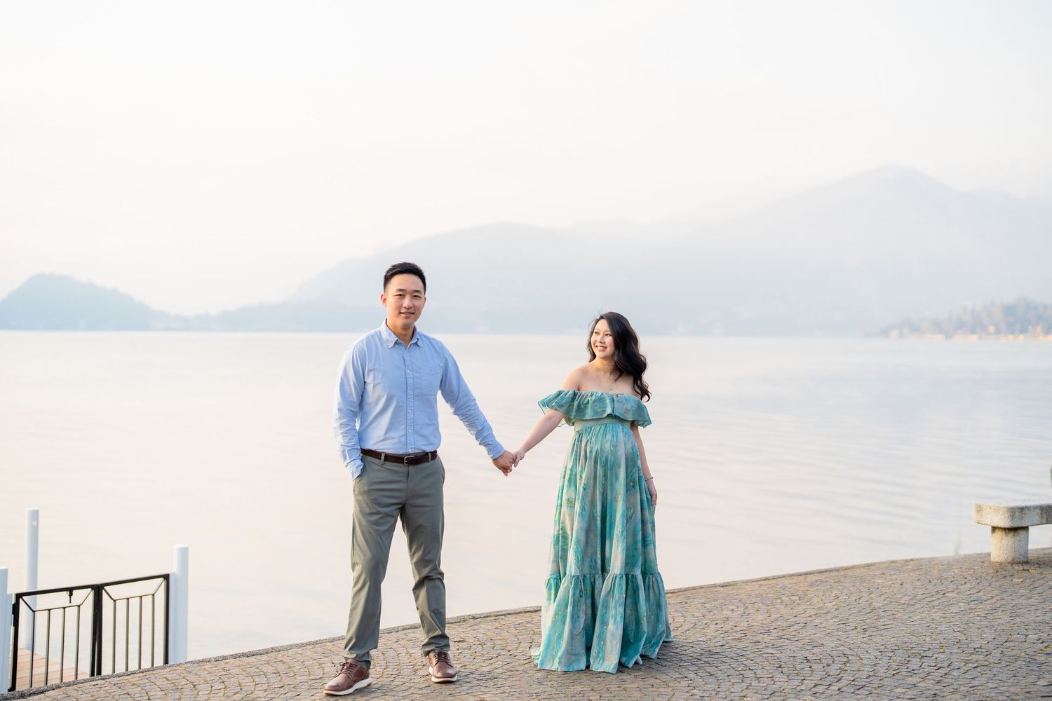 Couple holding hands by a lake with mountain backdrop, woman in teal dress, man in blue shirt, romantic outdoor photo.