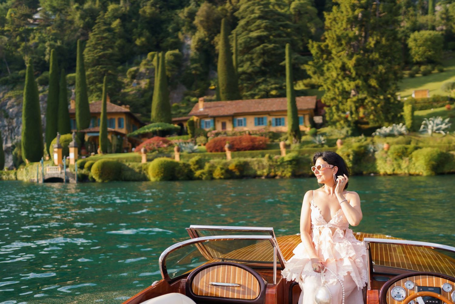 Elegant woman in pink gown on wooden boat at Lake Como, Italy, with lush gardens and villa in background.