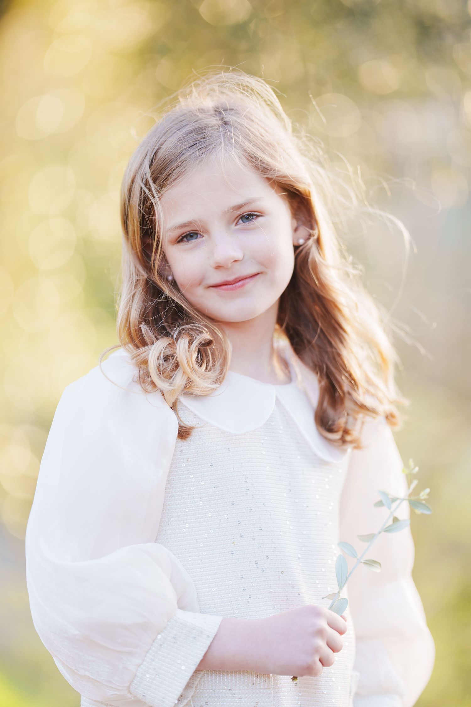 Young girl with long wavy hair holding a wildflower stem, wearing a white dress outdoors in soft golden light.