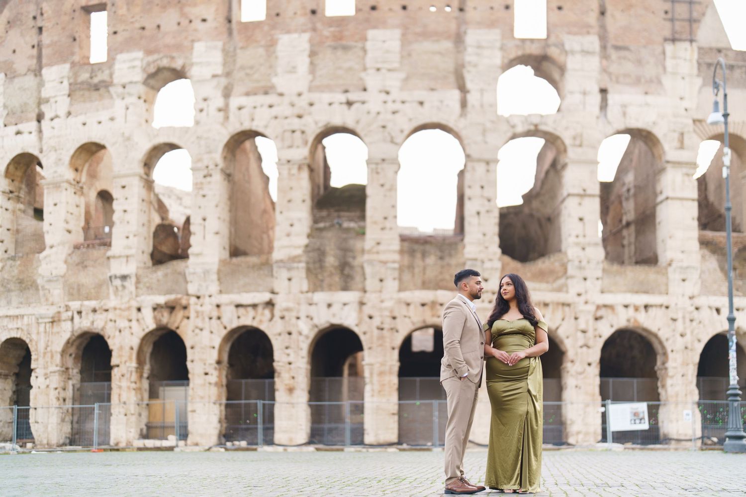 Couple posing in front of the Colosseum in Rome, woman in green dress, man in beige suit, maternity photo.