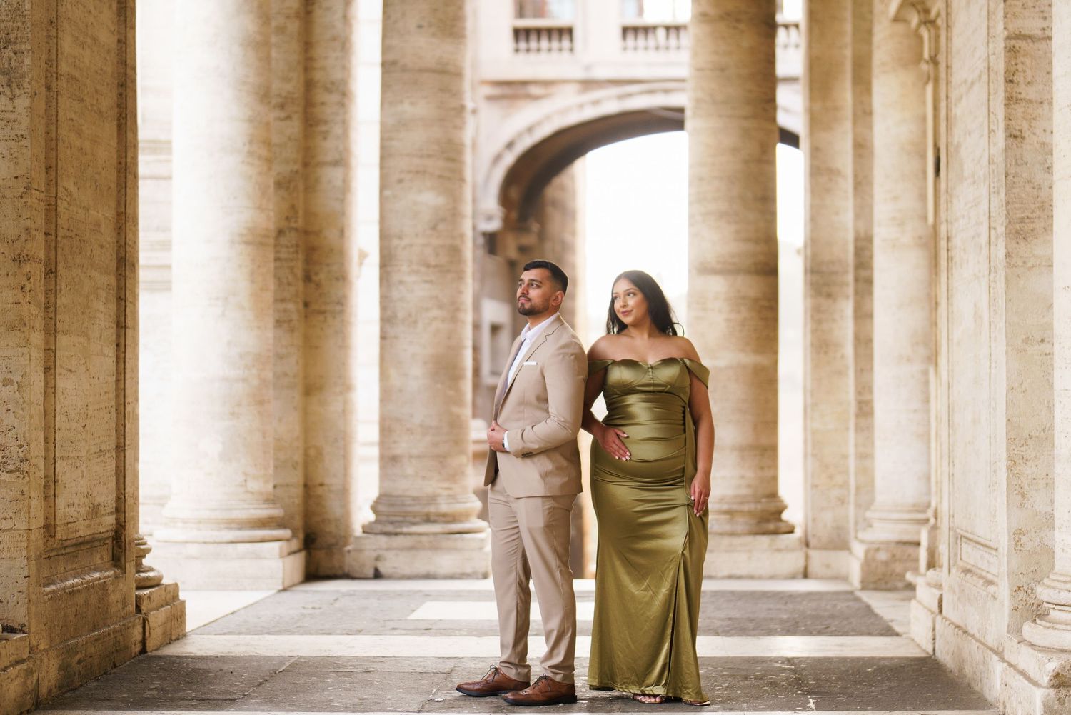 Elegant couple posing among grand stone columns in Rome, wearing formal attire in complementary gold and beige tones.