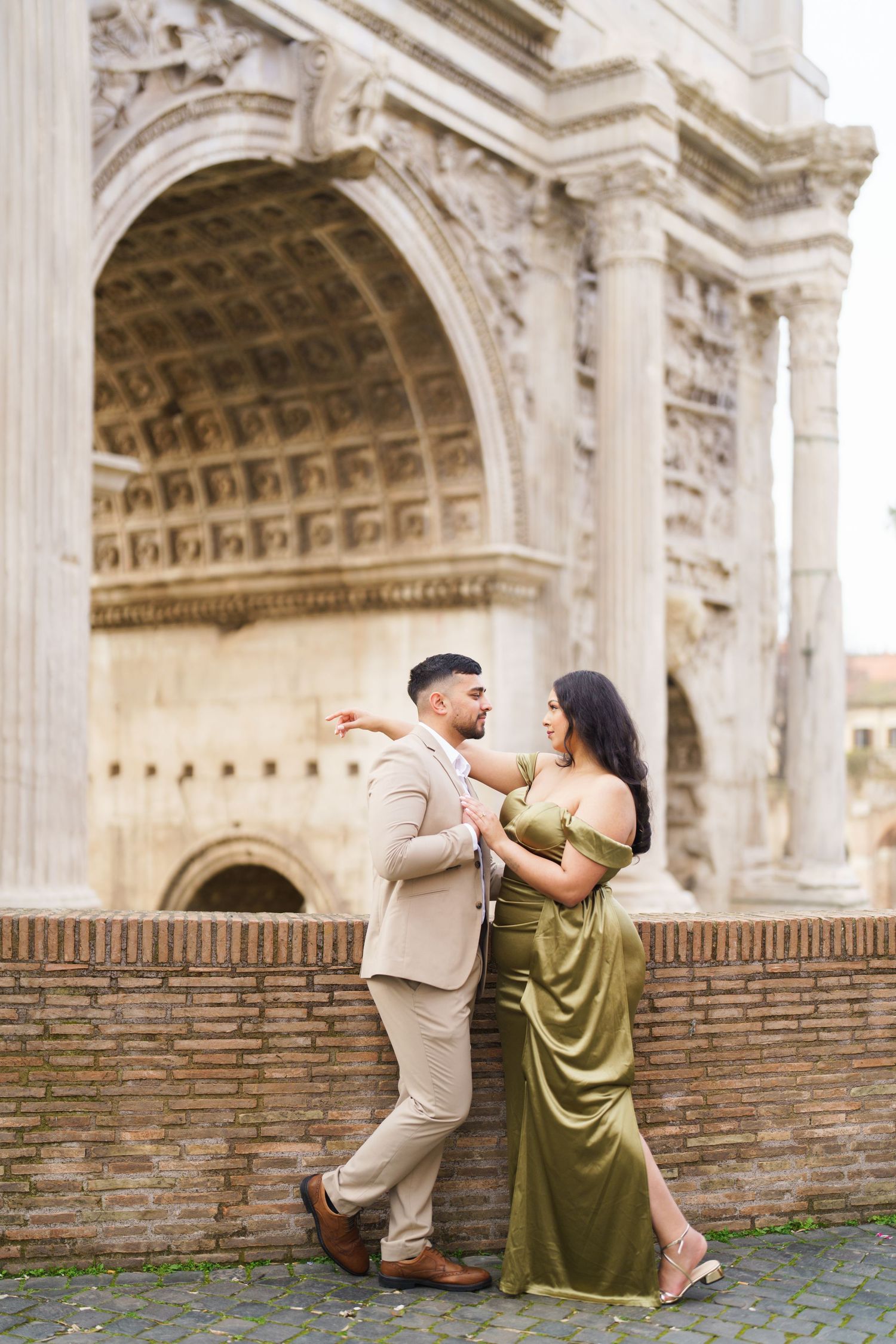 Couple posing romantically in front of an ancient Roman arch, man in beige suit, woman in elegant olive green gown.