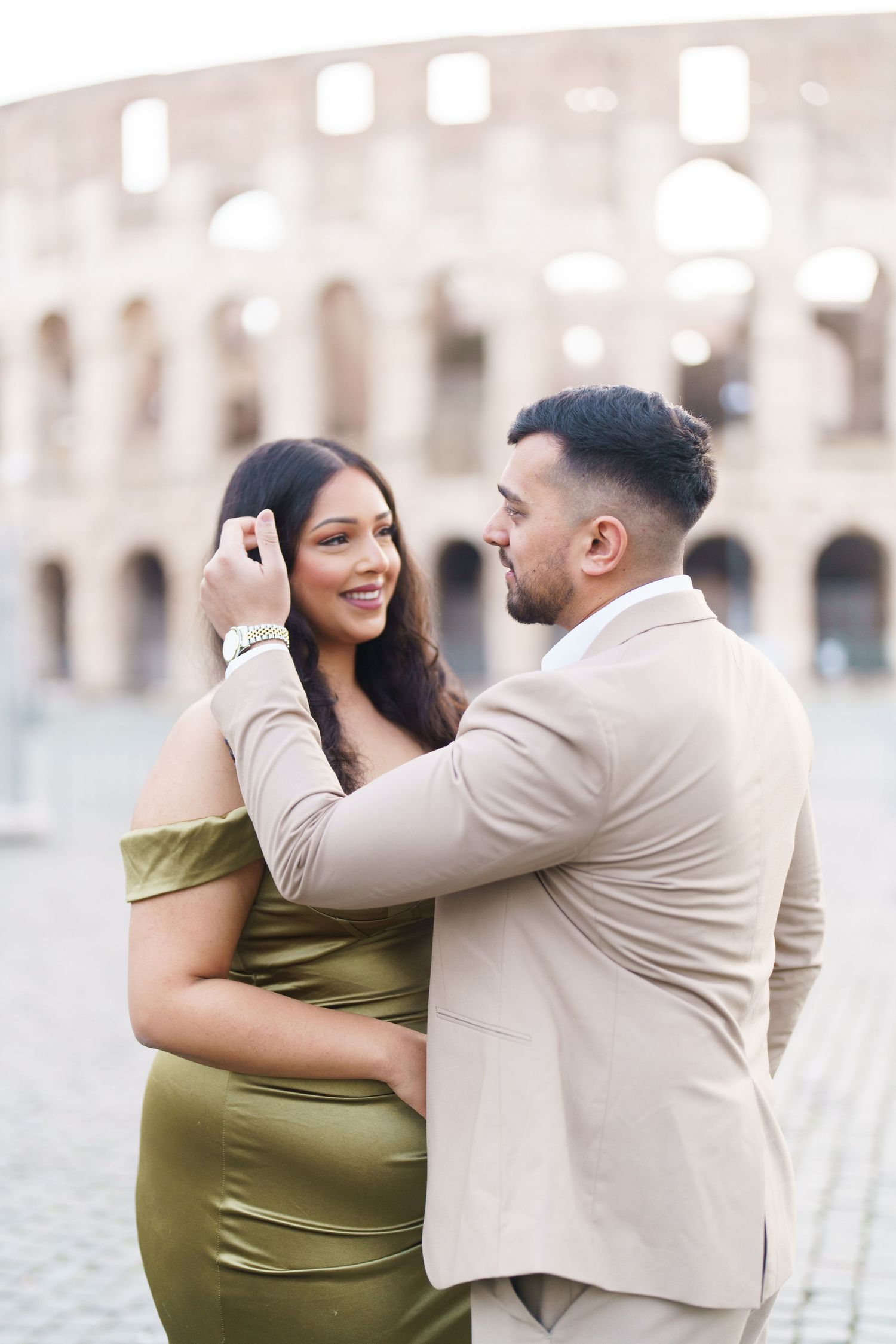 Couple embracing romantically in front of the Colosseum in Rome, woman in olive dress, man in beige suit.