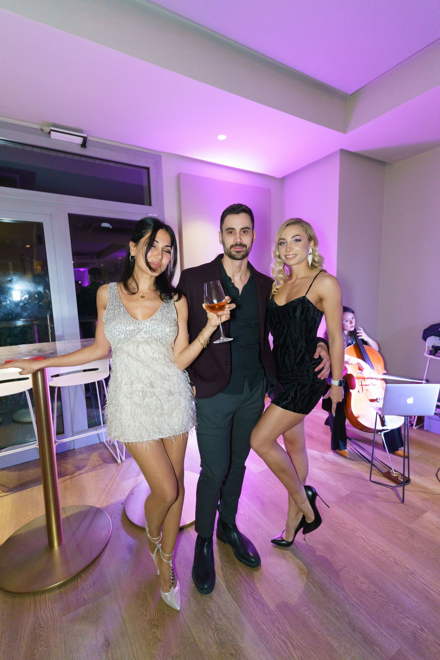 Three elegantly dressed party guests pose together at an upscale event with purple lighting and a cellist performing in the background.