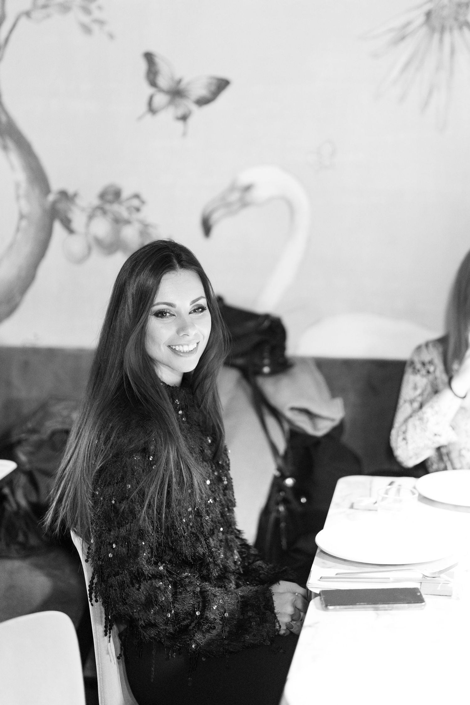 Black and white photo of a smiling woman with long hair sitting at a restaurant table with decorative bird mural behind her.
