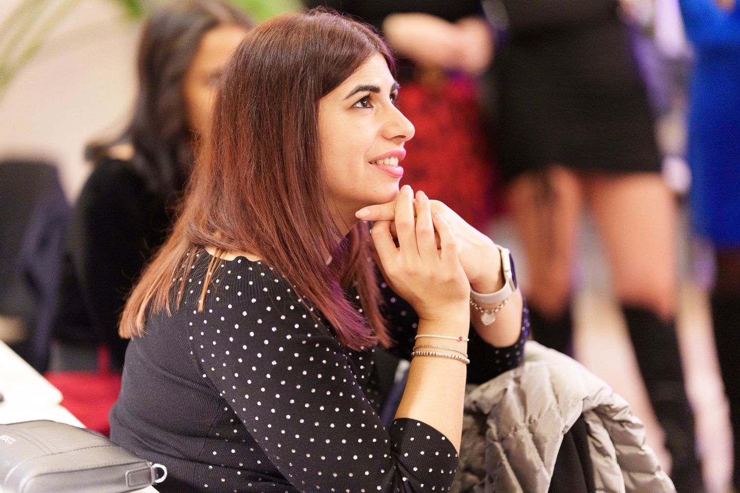 Young woman in polka dot blouse smiling thoughtfully with hands clasped, sitting at an indoor event.