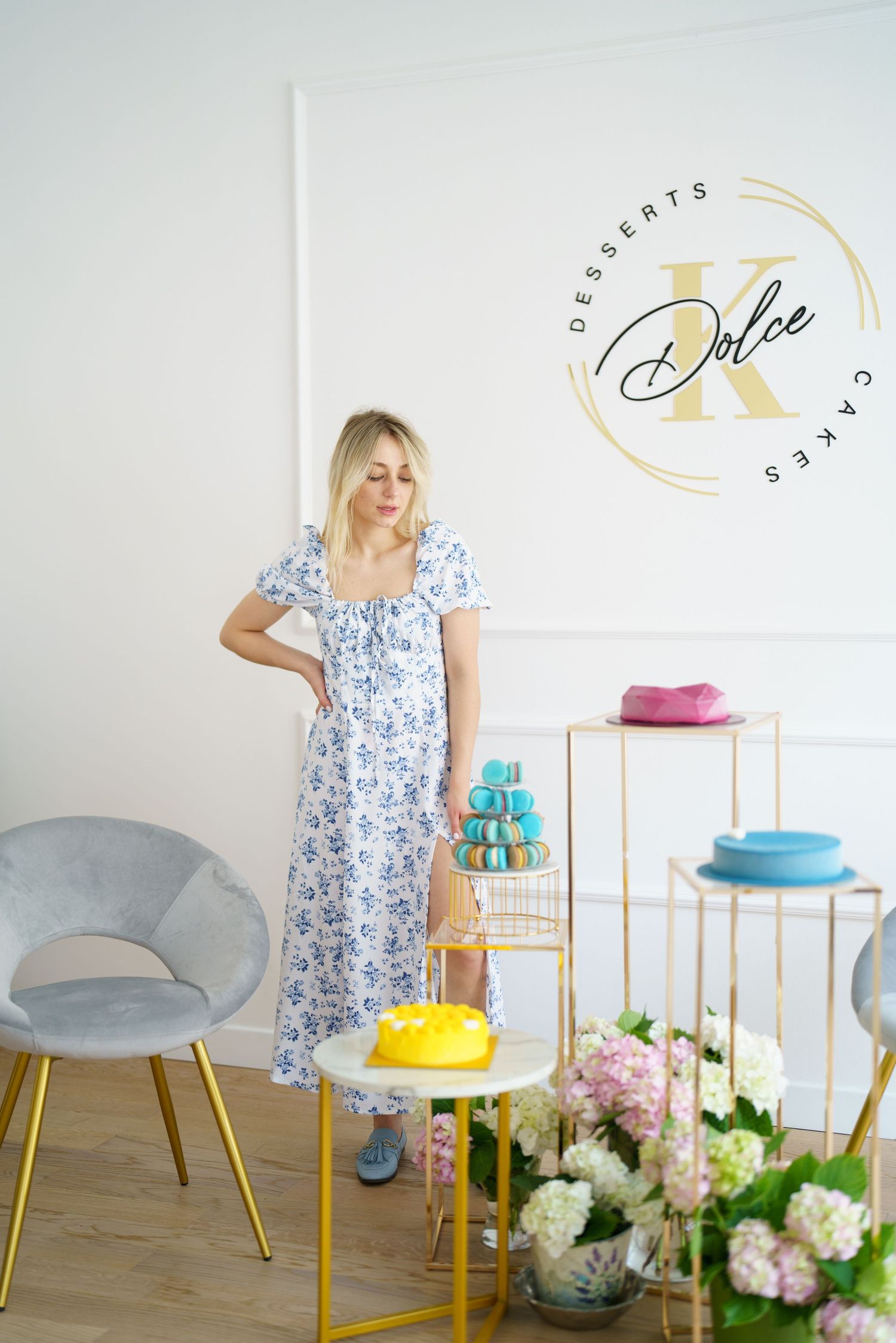 Blonde woman in floral dress poses in Dolce K desserts and cakes shop with macarons and yellow cake.