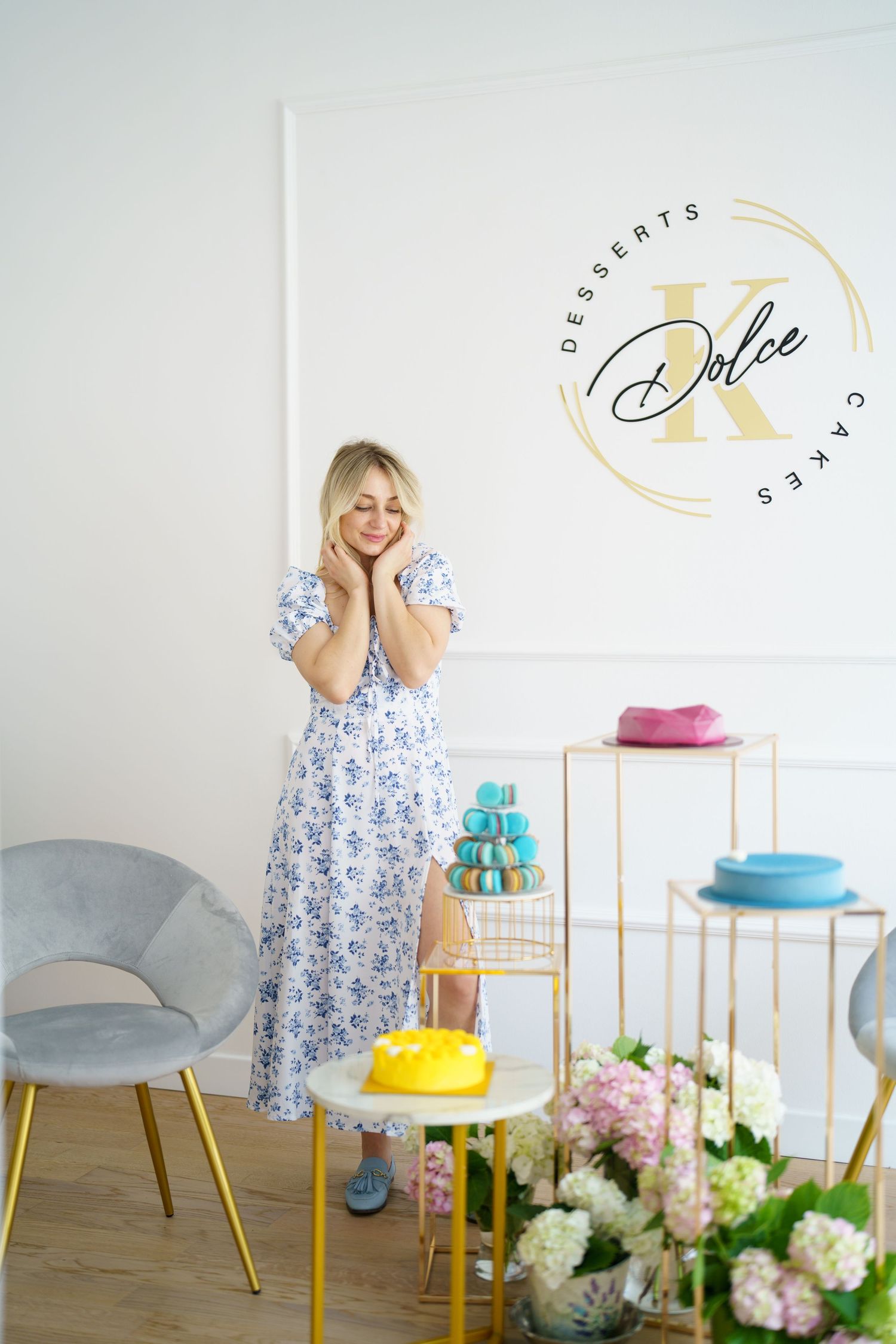 Woman in floral dress posing at Dolce X Desserts and Cakes shop with colorful macarons and cakes.