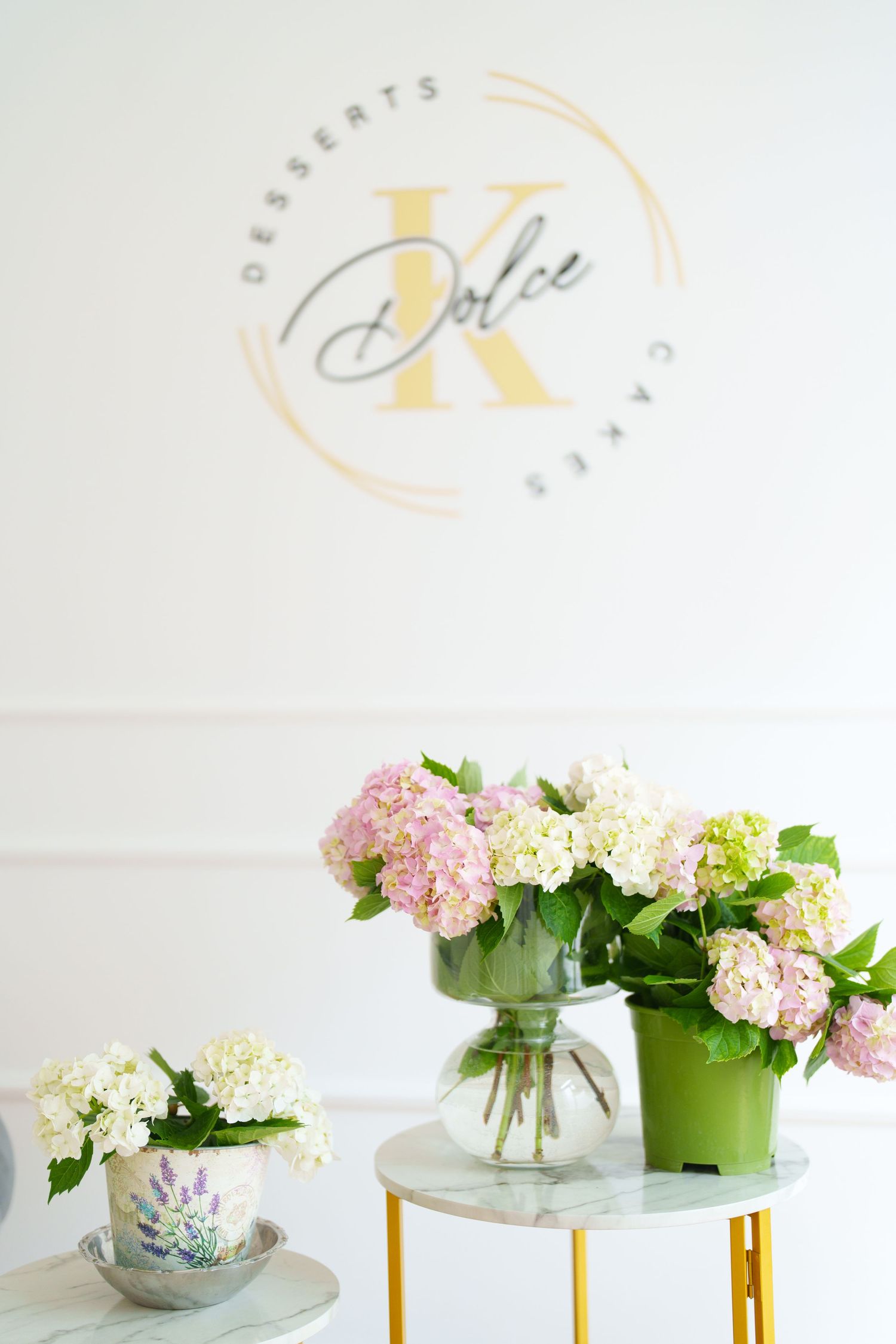 Elegant dessert shop display with pink hydrangeas in glass vase on marble table, K Dolce Cakes logo on white wall.