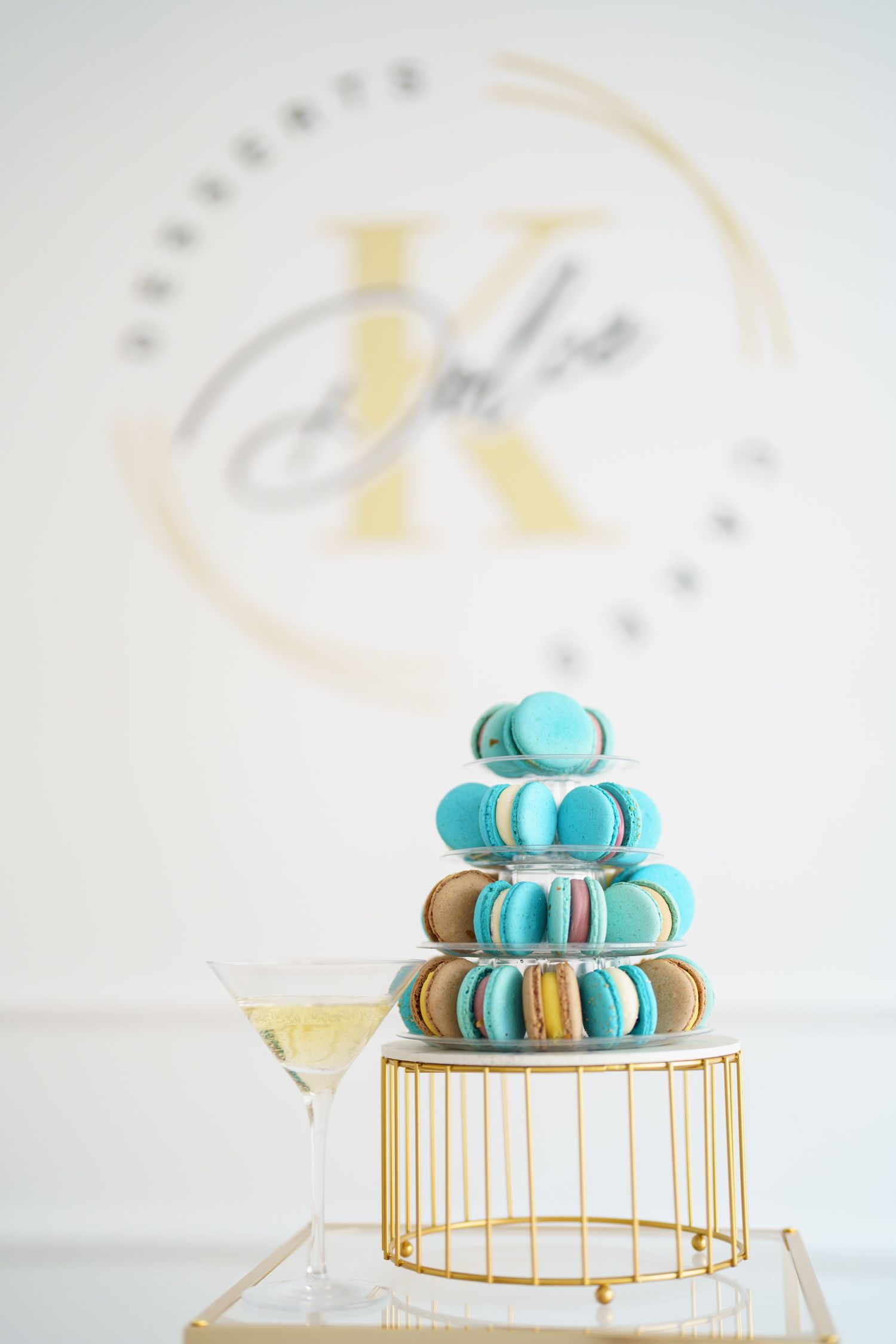 Elegant macaron tower display with turquoise and brown macarons on gold stand beside champagne glass.