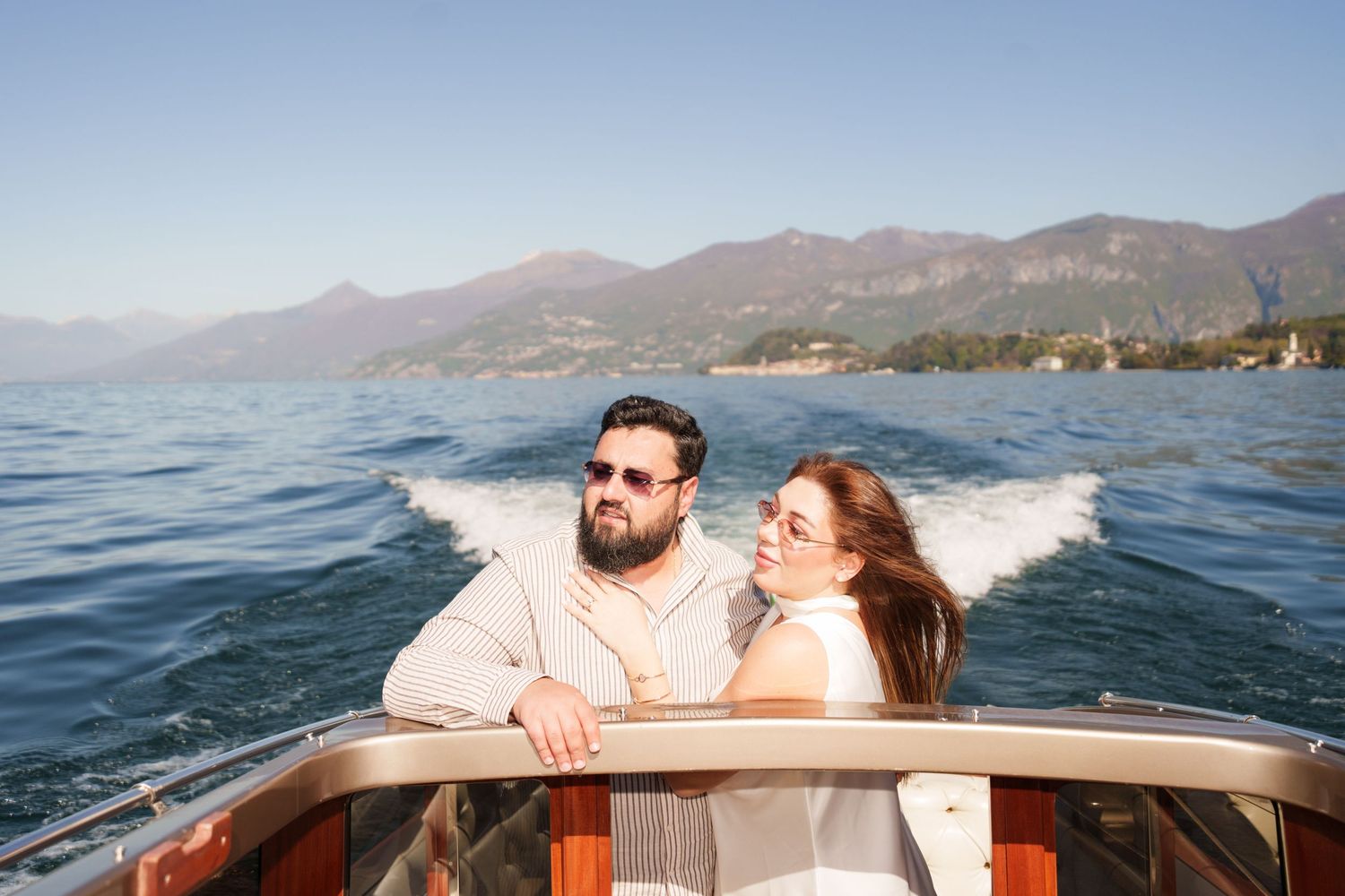 Couple enjoying a scenic boat ride on a mountain lake, woman leaning on man in sunglasses, clear blue sky.