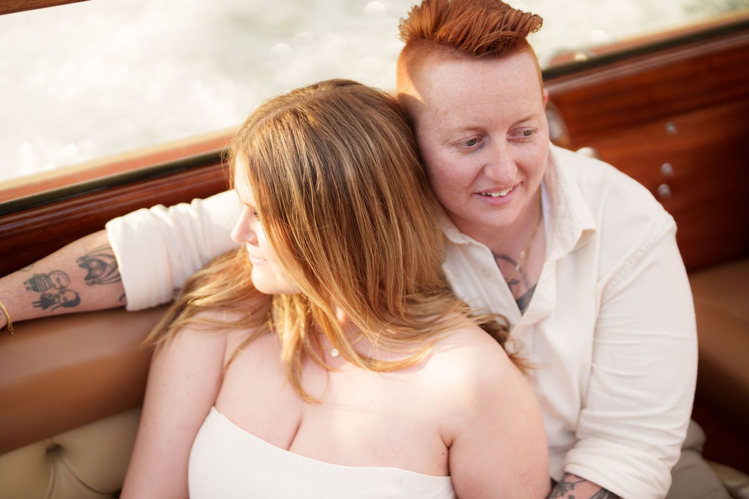 Two women share a tender moment seated together on a vintage leather car seat, one with red hair and tattoos.