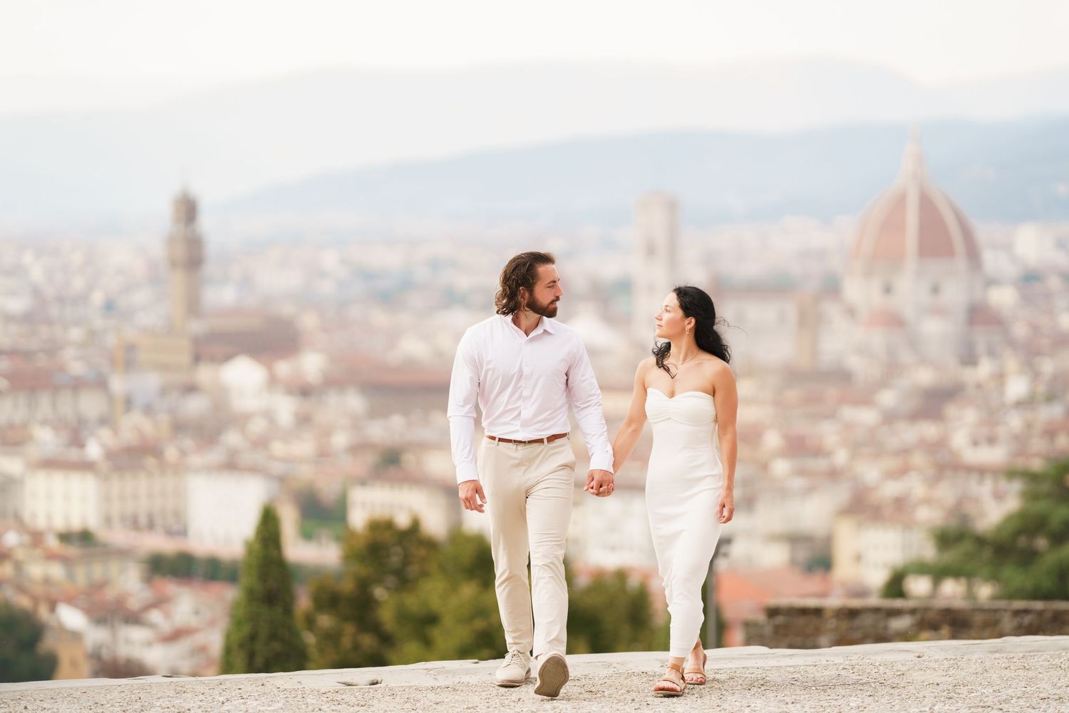 A couple walks along a scenic overlook with Florence's iconic Duomo cathedral and cityscape in the background.