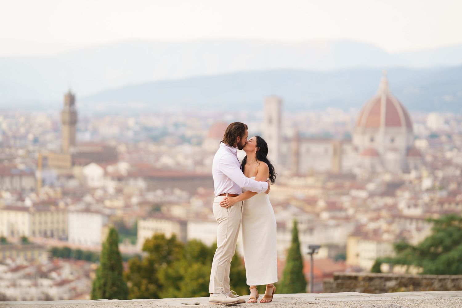 A romantic couple embraces while overlooking the historic skyline of Florence, Italy with the Duomo cathedral in the background.