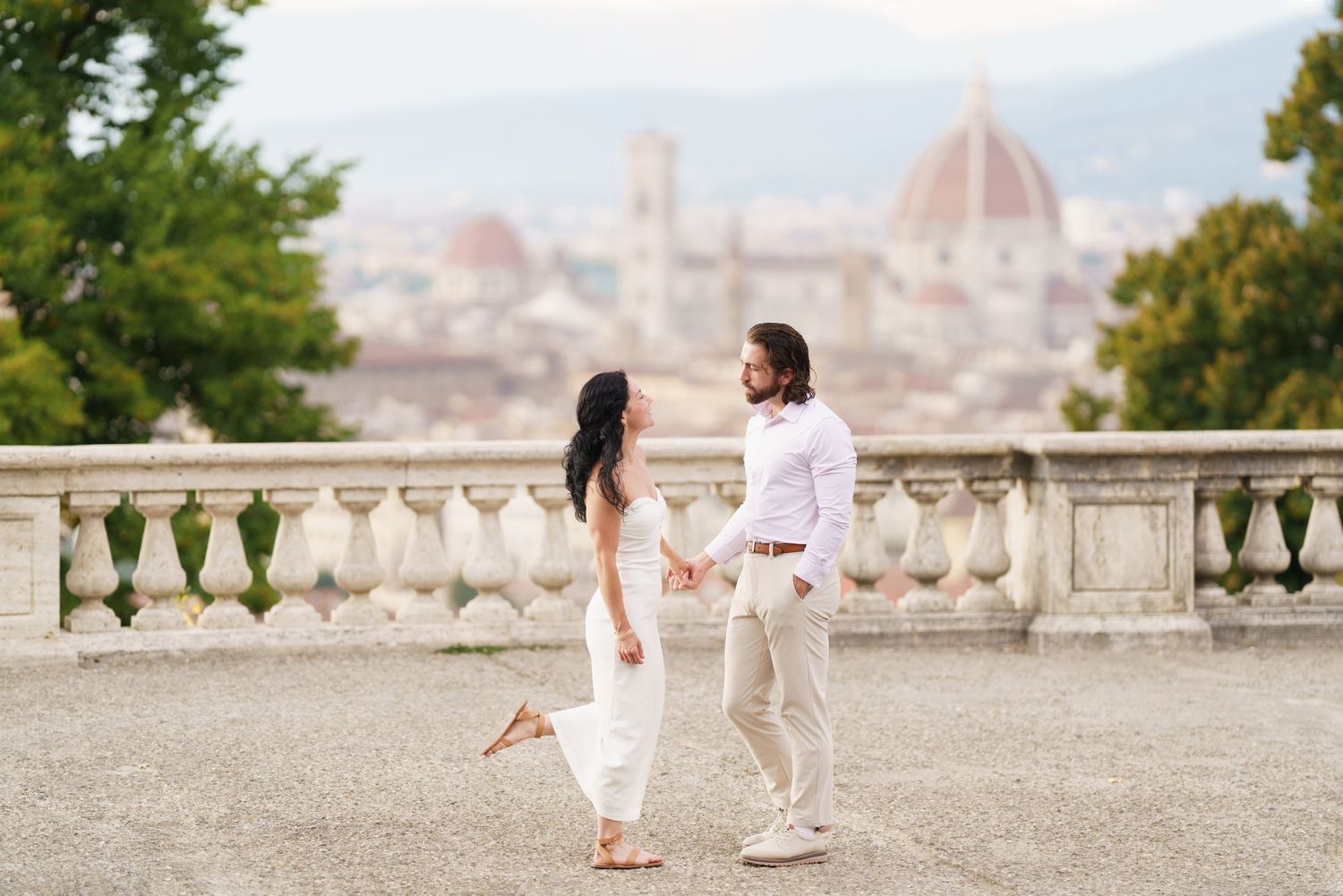 A romantic couple in white outfits poses on a terrace overlooking Florence, Italy with the Duomo cathedral visible in background.