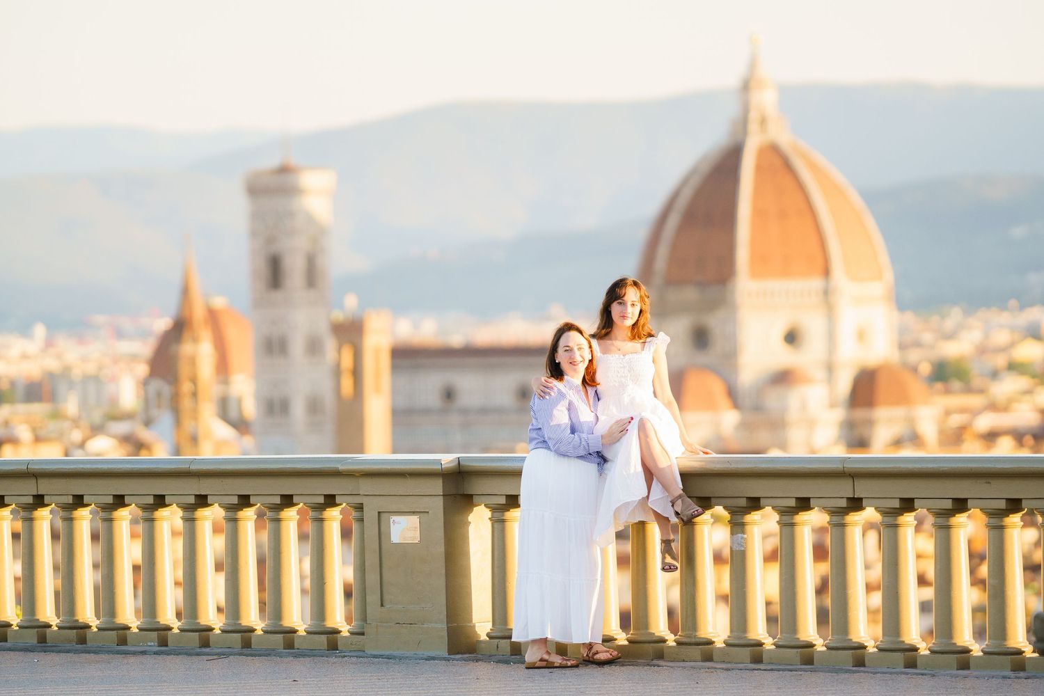 A couple in white outfits embrace at Piazzale Michelangelo overlooking Florence's Duomo cathedral during sunset.