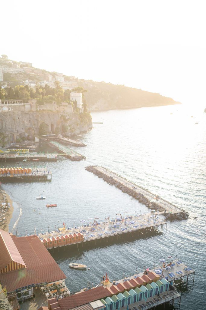 Scenic view of a coastal marina with boats and buildings nestled against cliffs at sunset.