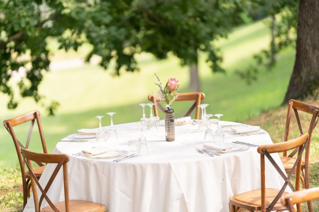 Outdoor wedding reception tables set with white linens and wooden cross-back chairs under trees.