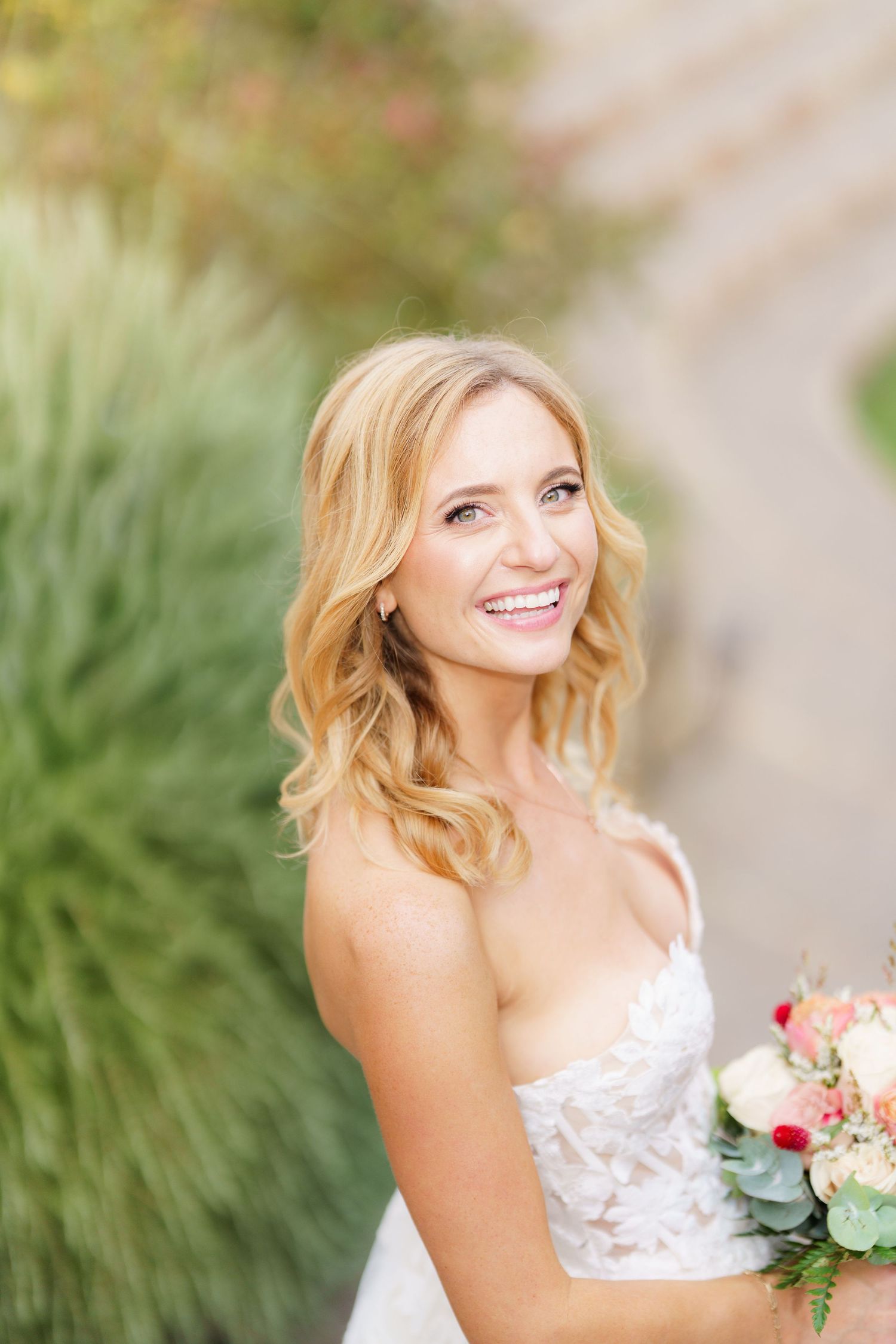 Bride in white lace dress poses outdoors with natural greenery background.