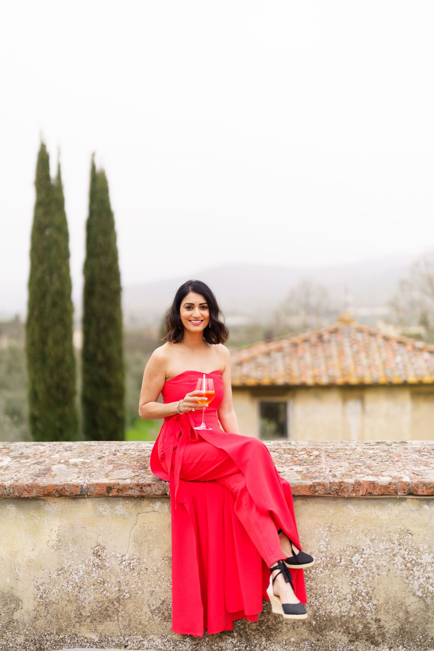 Person in a vibrant red dress sits on stone wall with cypress trees and countryside views.