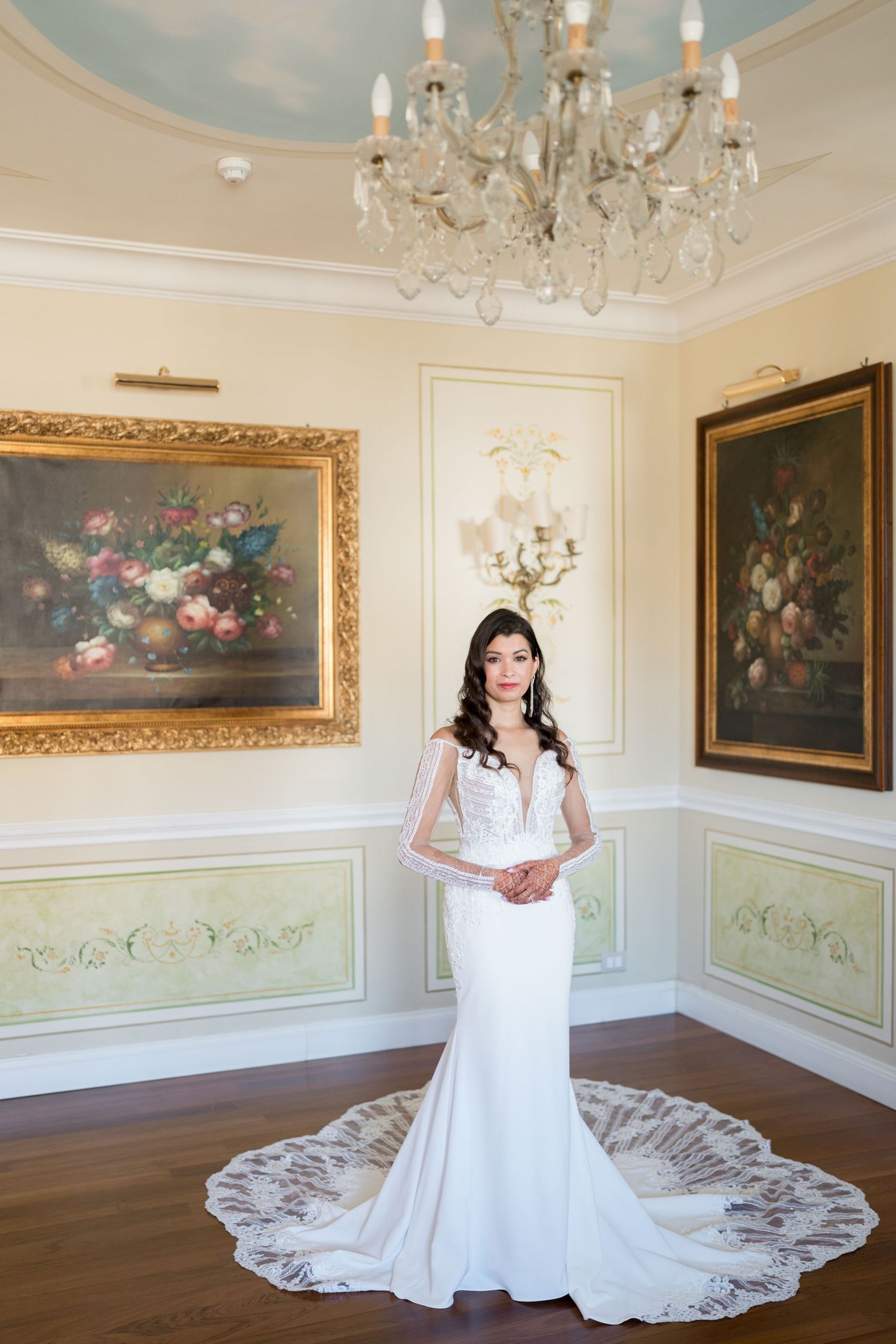 A wedding dress with intricate lace train displayed in an elegant room with chandeliers.