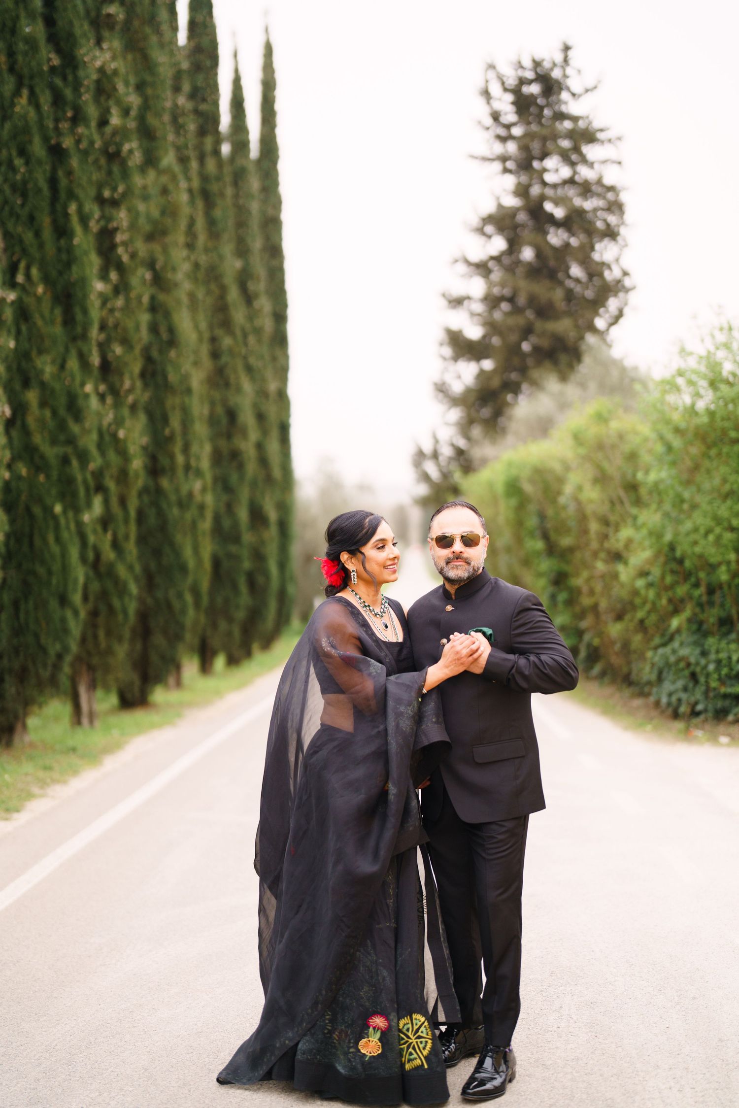 Couple in formal black attire dancing on cypress-lined path.