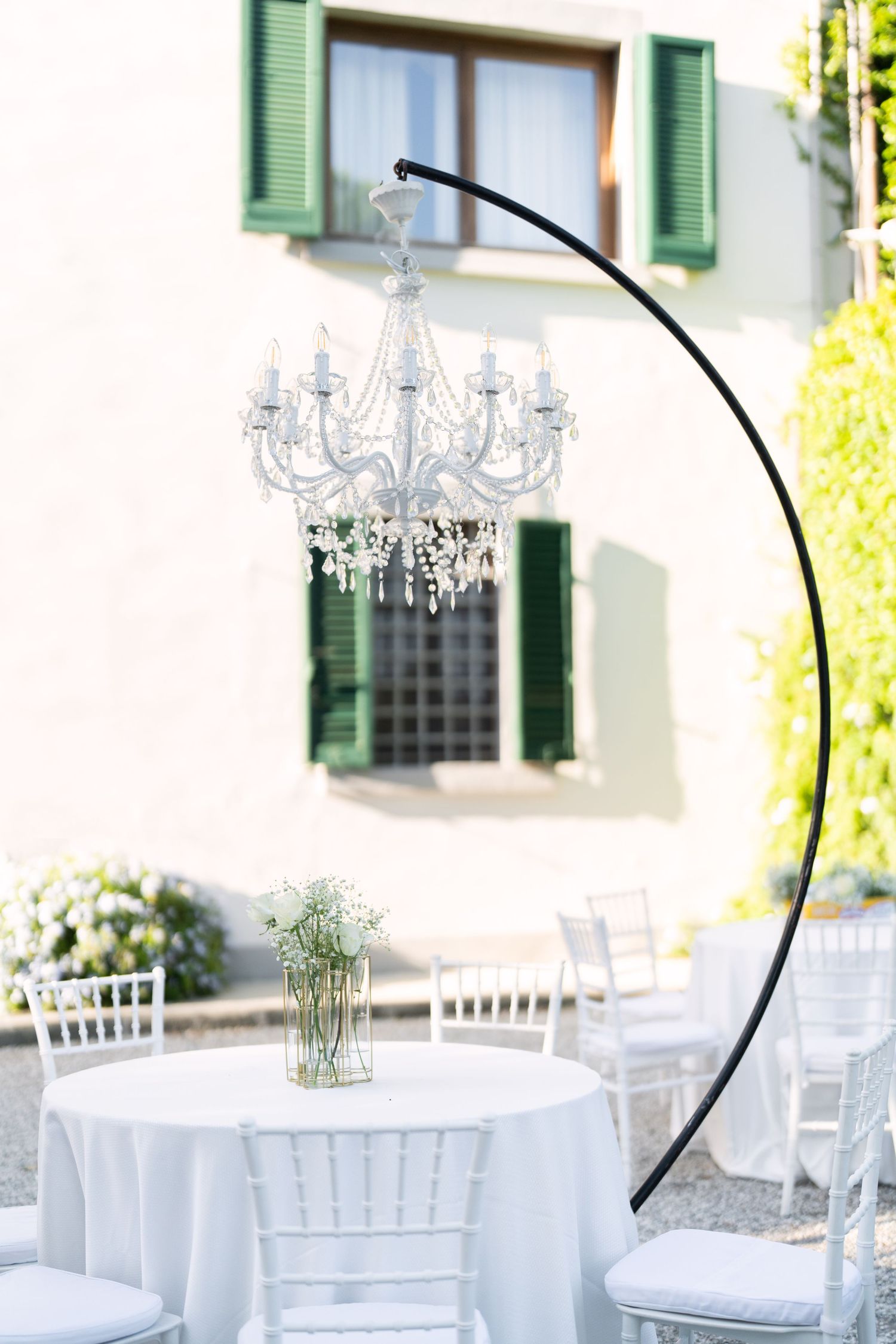 Crystal chandelier suspended from black arch with white table and chairs against building with green shutters.