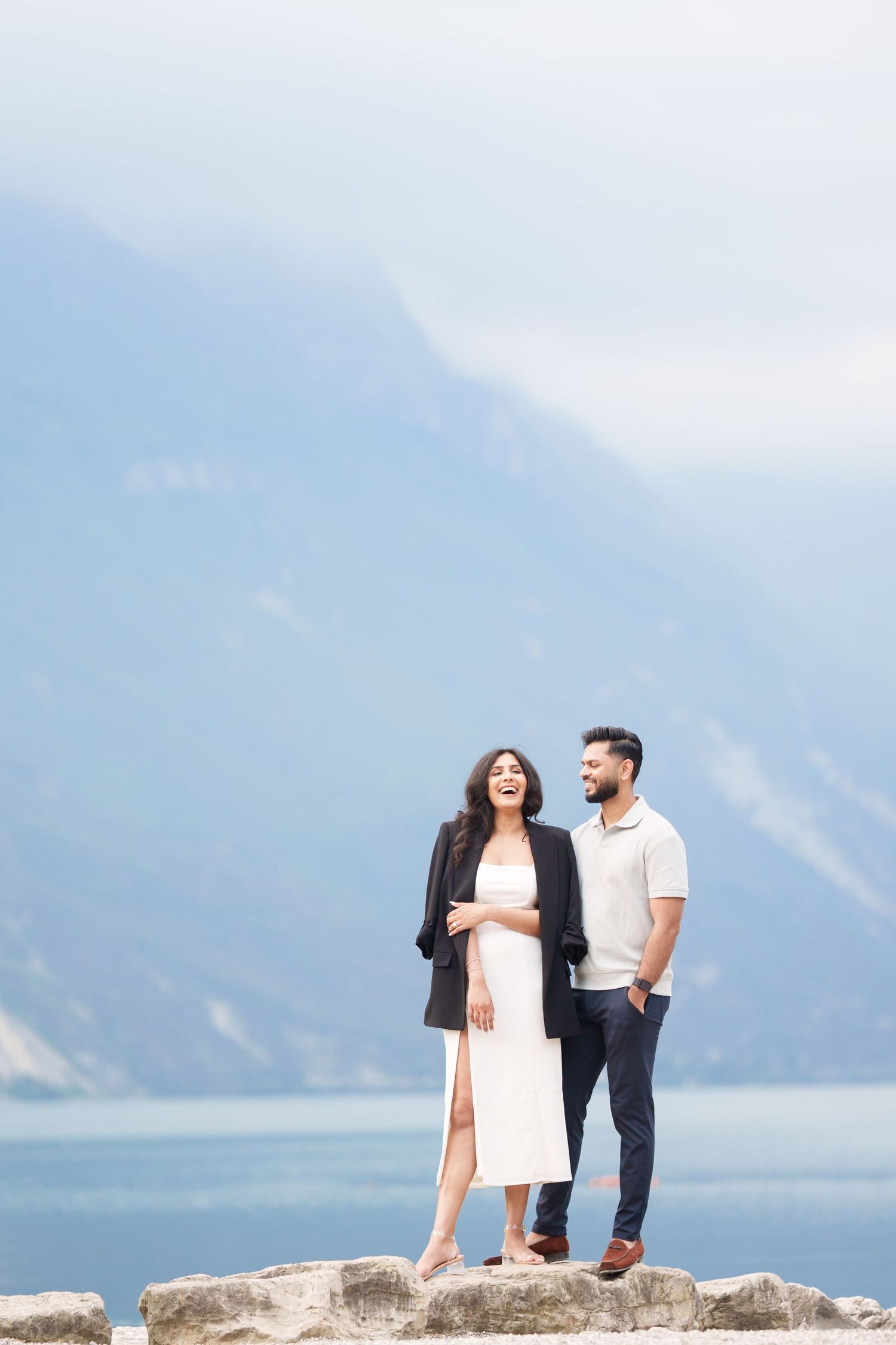 Couple sharing a joyful moment together on rocky cliff overlooking a misty mountain lake landscape.