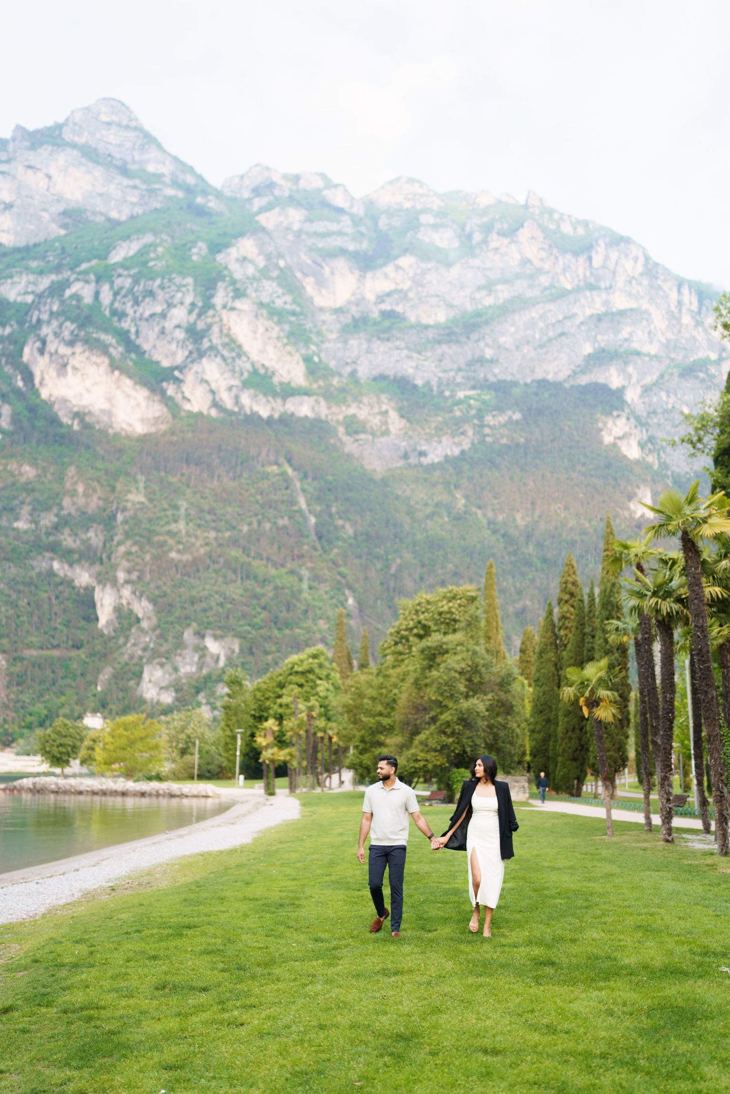 Two people stroll along a lakeside path with dramatic mountain peaks and palm trees in Lake Garda, Italy.