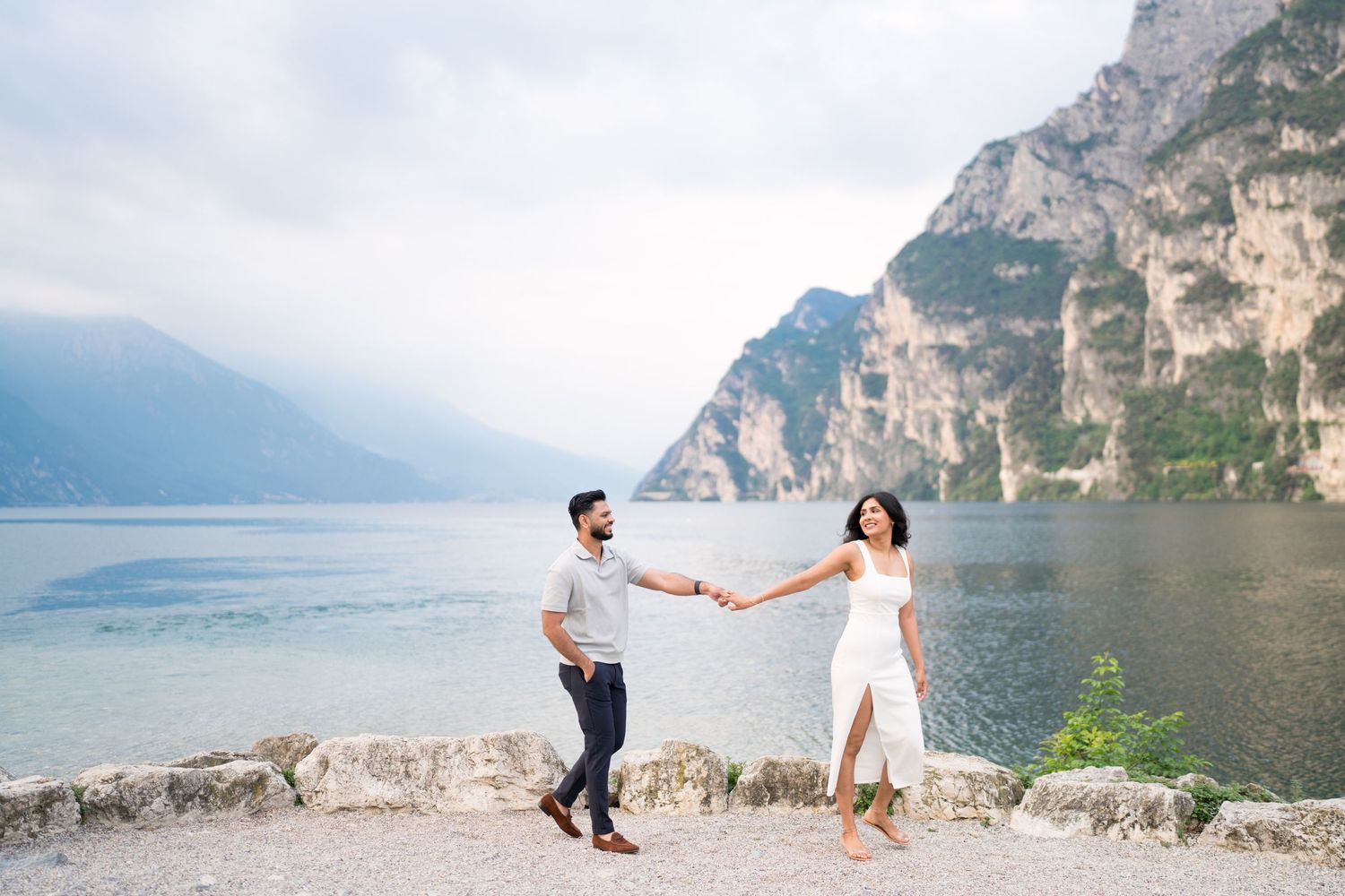Couple in white outfits walking along scenic mountain lake shore with dramatic cliffs in the background.